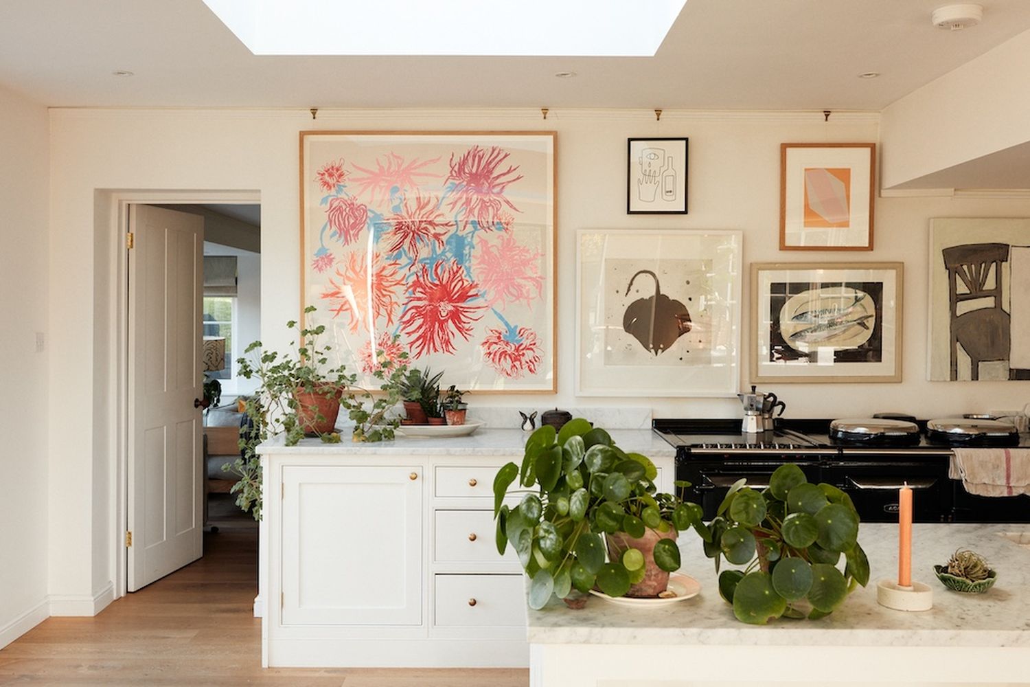 A kitchen with a white countertop and a black stove. There are potted plants on the countertop and a large potted plant in a white pot. The walls are adorned with three framed pictures. [G]