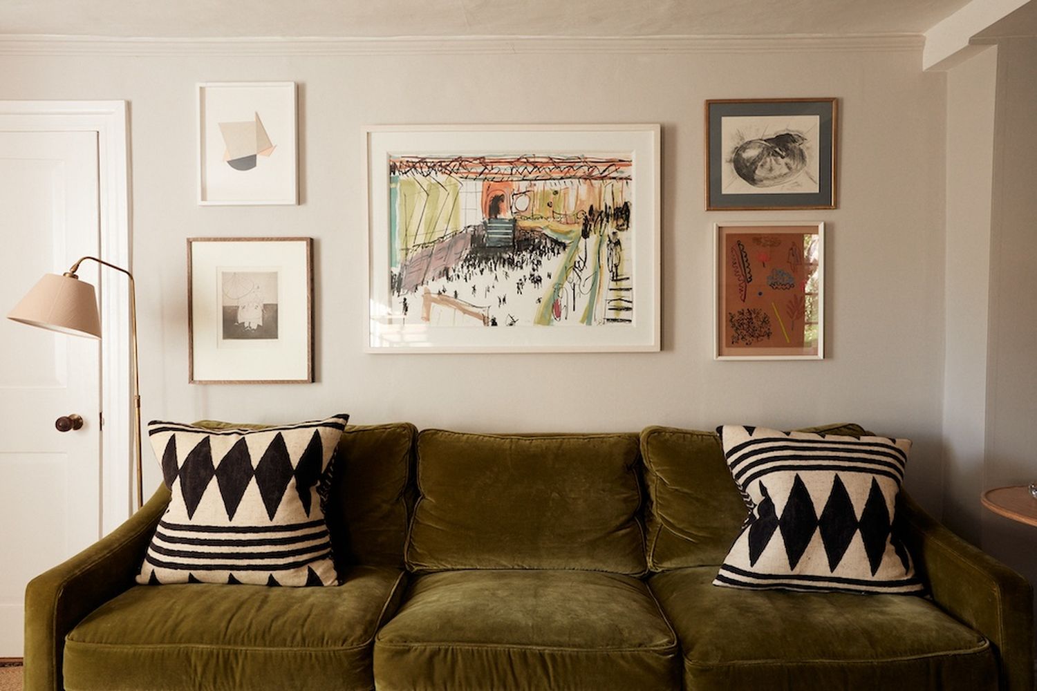 A living room with a green couch adorned with black and white striped pillows. The wall behind the couch is decorated with various framed artworks. [G]