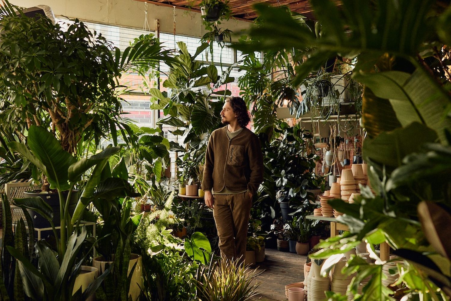 A young man stands in a greenhouse filled with various plants, surrounded by pots and hanging plants. [G]