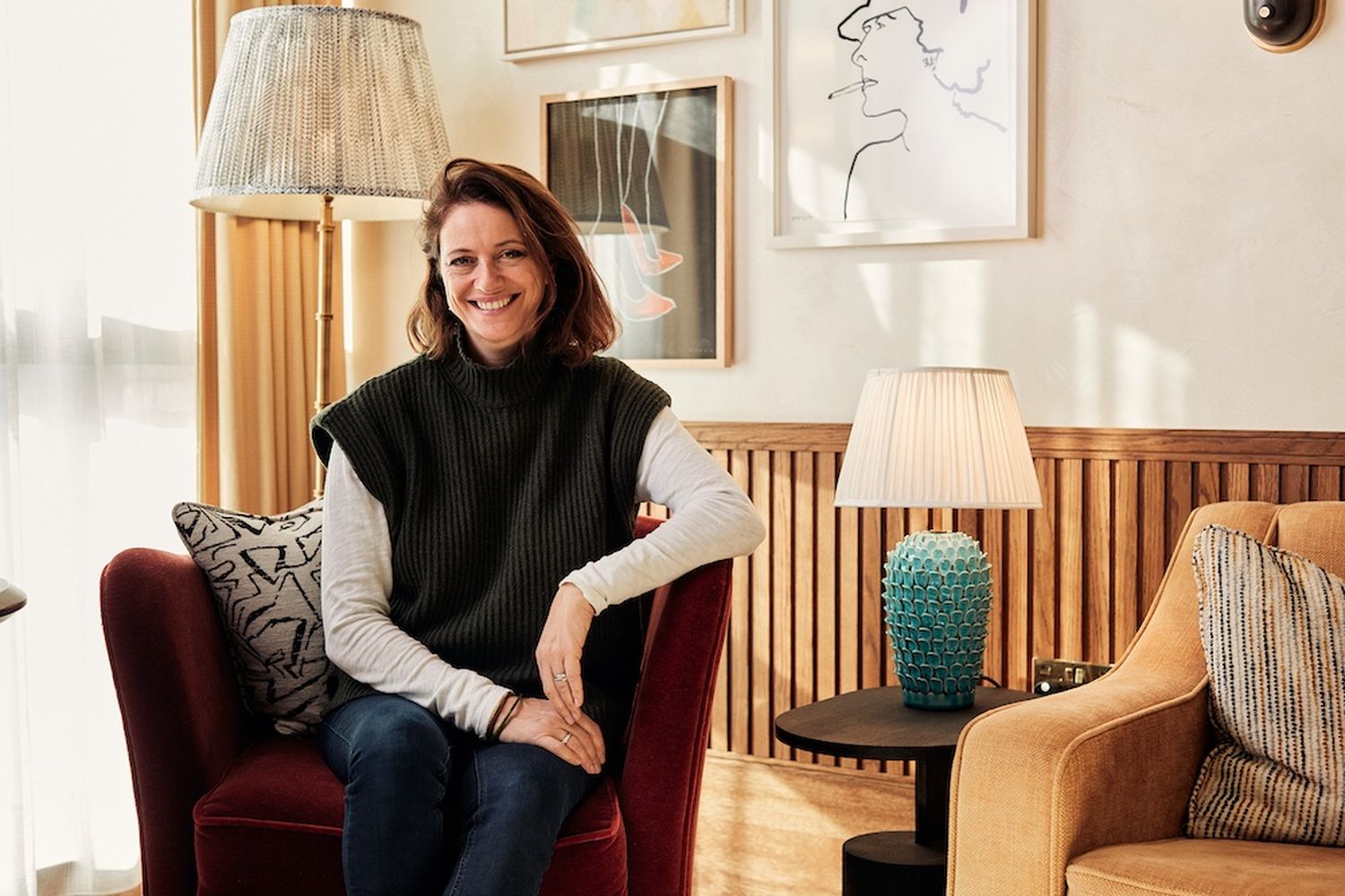 A woman in a black turtleneck sweater and blue jeans, sitting on a red armchair, smiles at the camera. The room is adorned with paintings and a blue vase on a side table. The wooden wall behind her adds warmth to the scene. [G]
