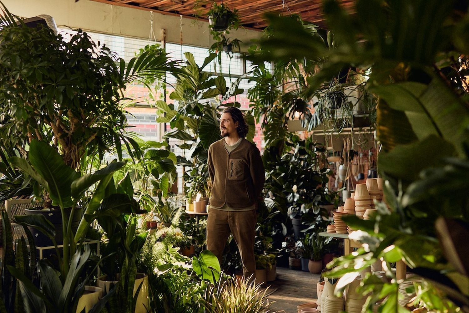 A man in a brown sweater and pants stands in a greenhouse filled with various plants. [G]