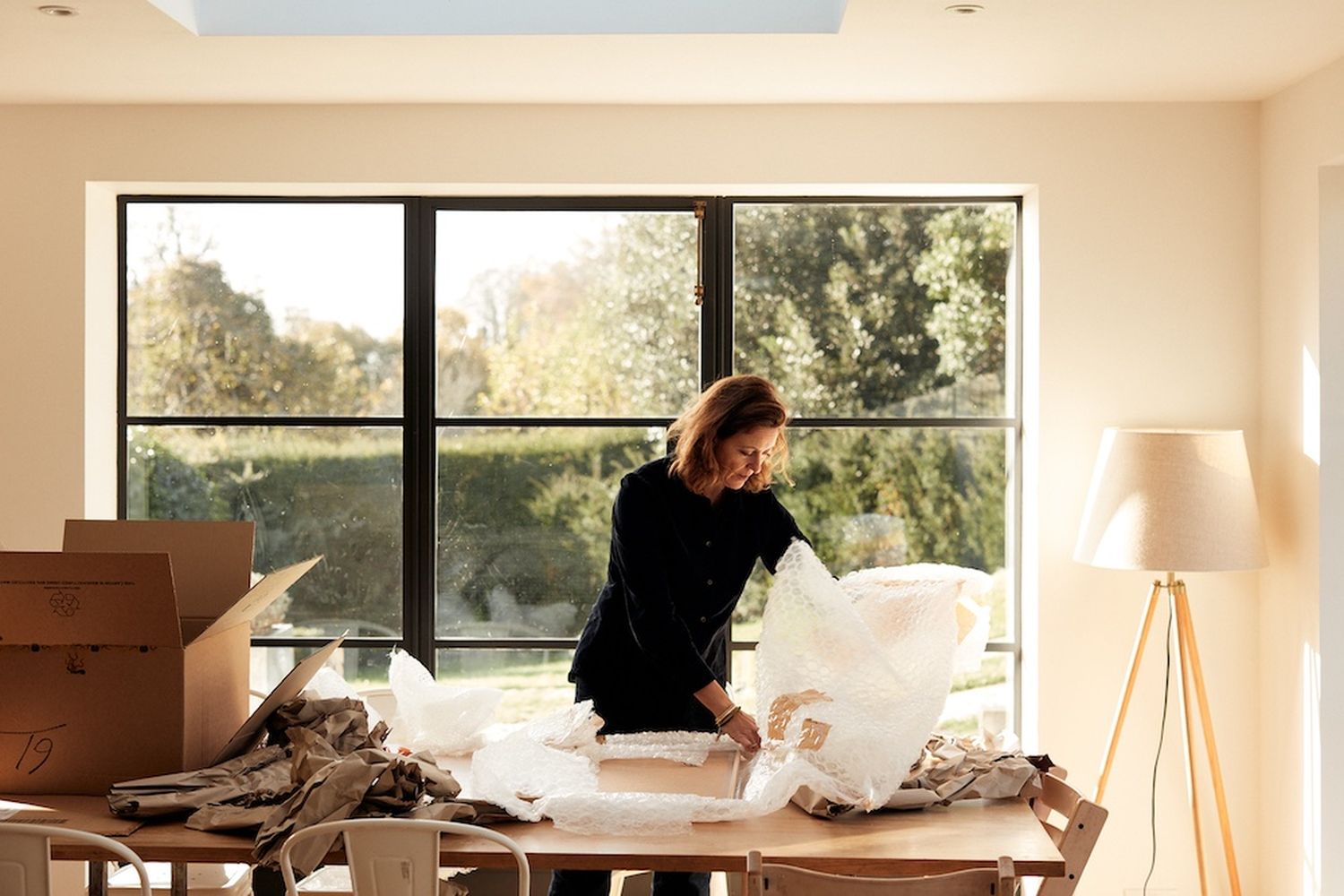 A woman in a black shirt is working at a table in a room with large windows. She is holding a white plastic bag and appears to be sorting or arranging it. The table is covered with a pile of papers and other materials. The room has a wooden floor and a large window that offers a view of trees outside. [G]