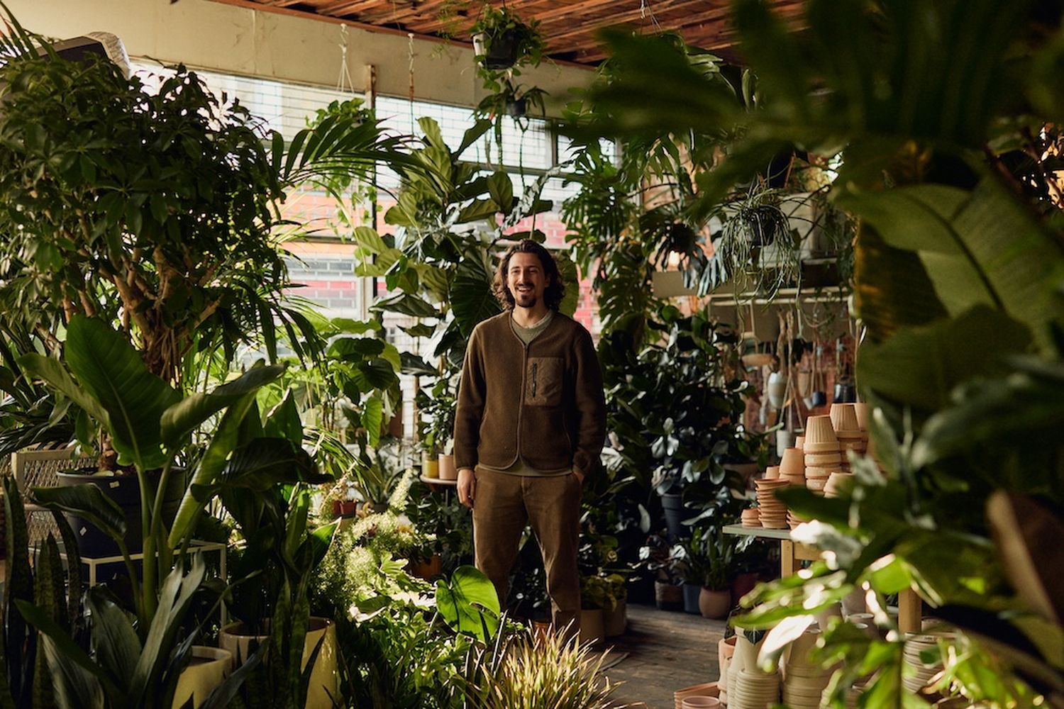 A man in a brown hoodie stands in a greenhouse filled with various plants. [G]