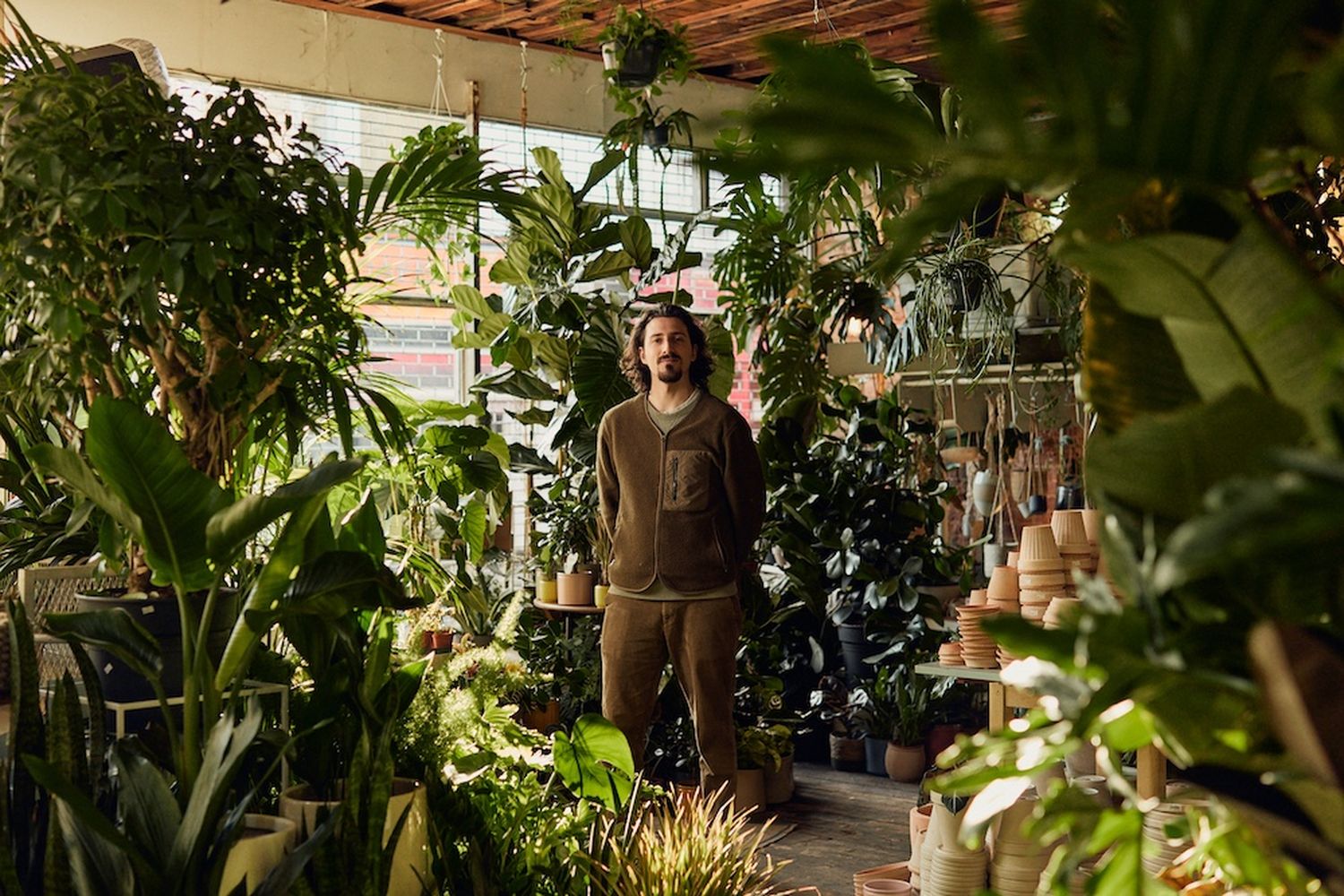 A man in a brown sweater stands in a greenhouse filled with various plants. [G]