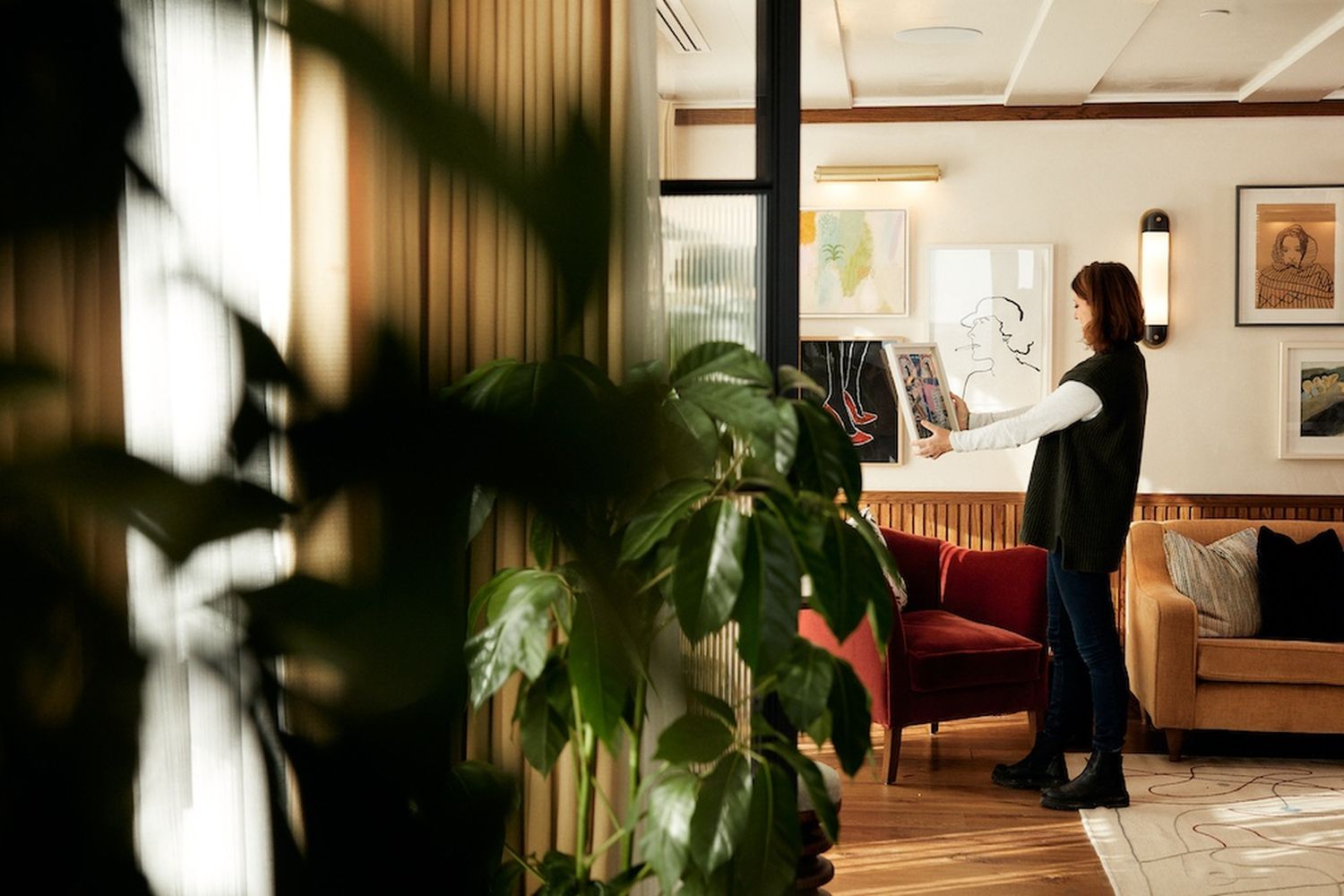 A woman in a black top and jeans is pointing at a map on a wall. She is standing in a room with a red armchair, a potted plant, and a window. [G]