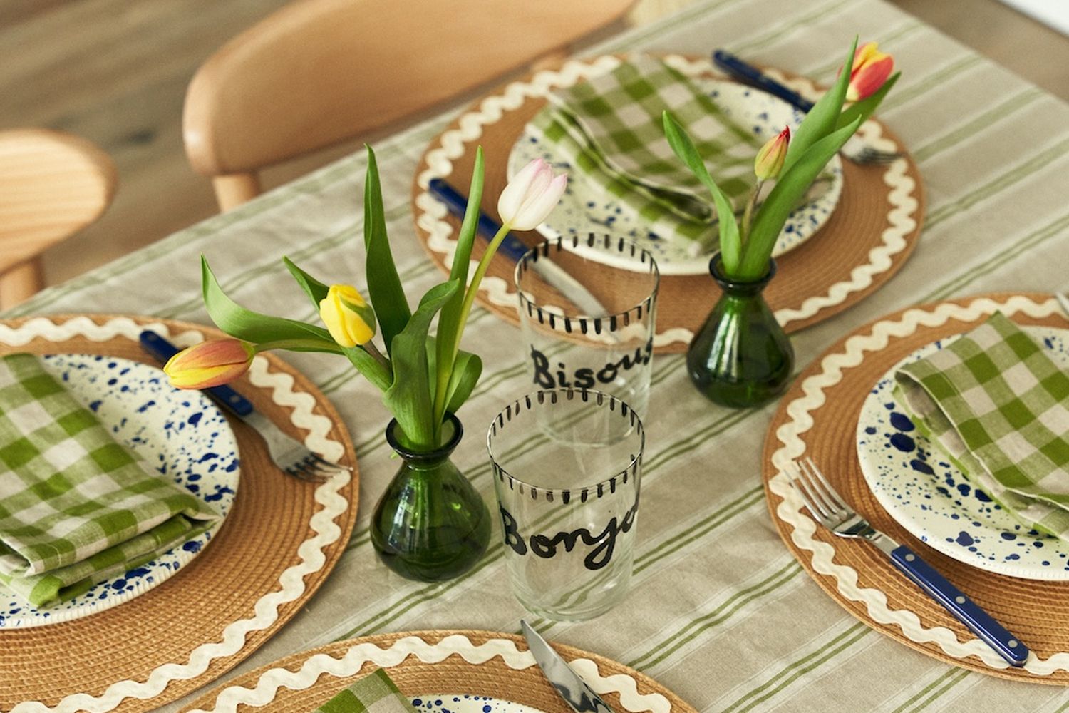 A table with four places with a green and white striped table cloth, natural circular placemats with white rickrack around the edges, blue splatter plates and green gingham serviettes.