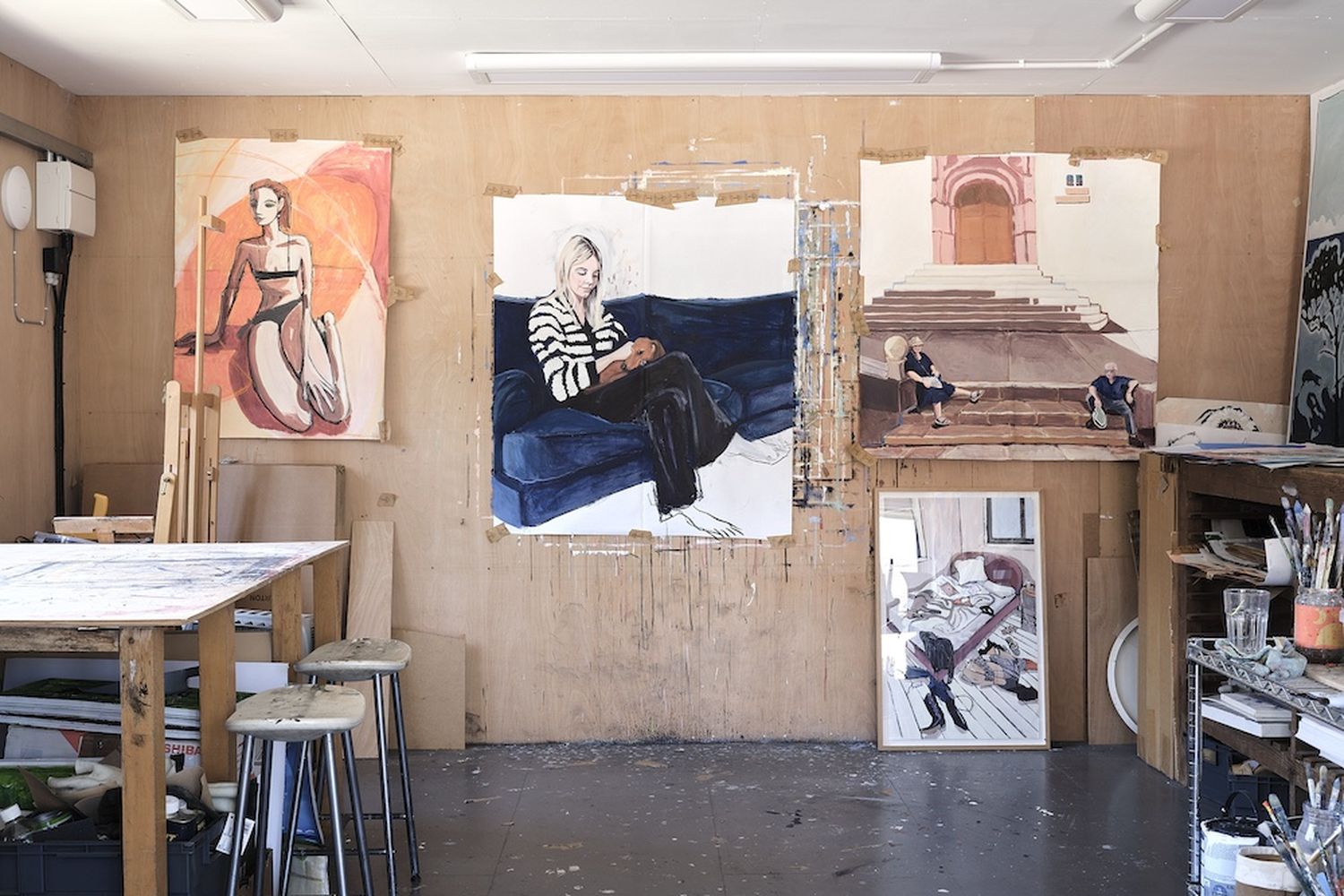 A woman in a striped sweater sits on a blue sofa, surrounded by various paintings of women in different poses. The room has a wooden floor and a table with stools. [G]