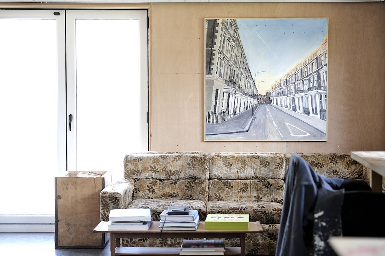 A living room with a floral couch, a coffee table with books, and a painting of a street scene. [G]