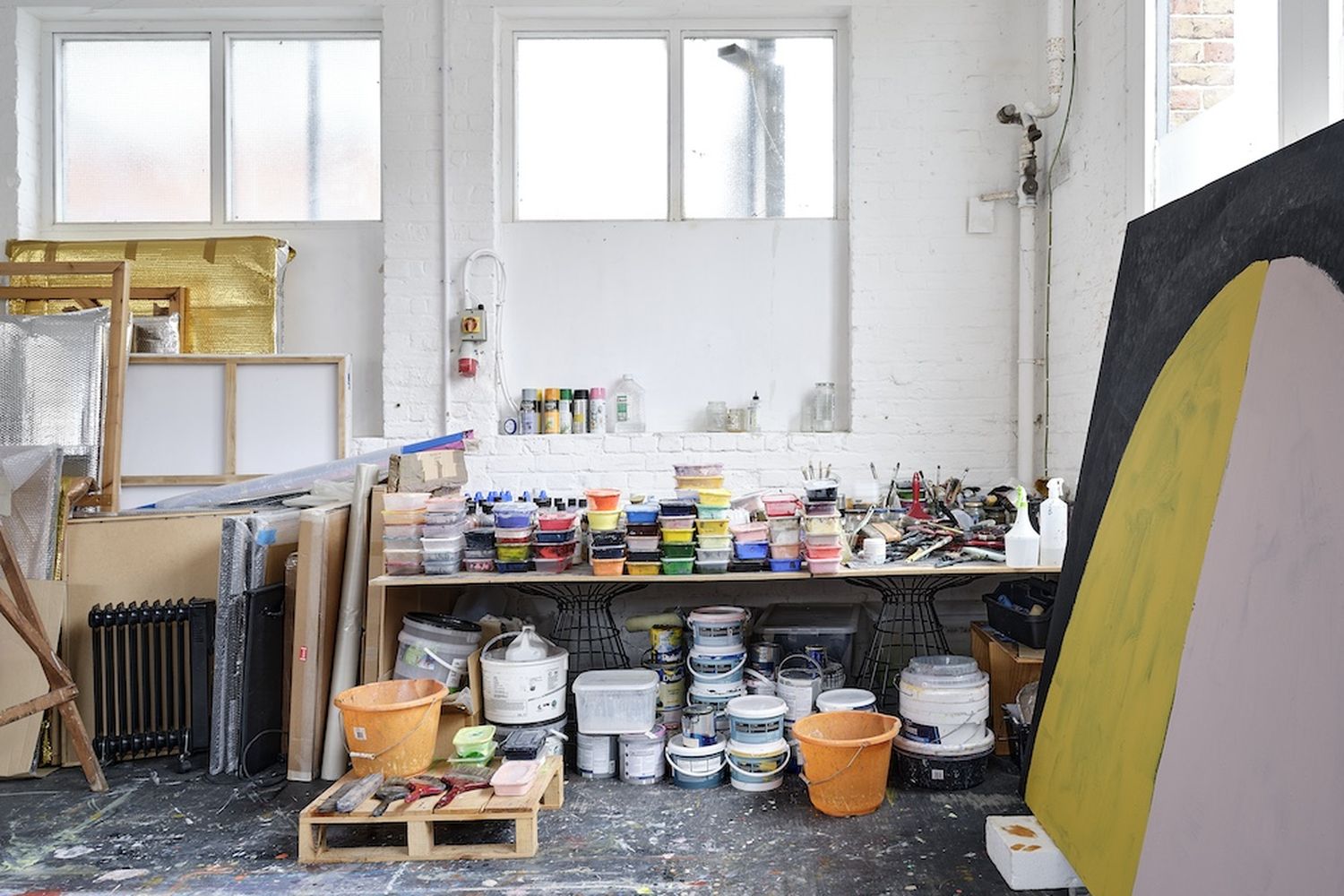 A cluttered studio with a large window, a wooden shelf filled with paint, and various paintbrushes and buckets. [G]