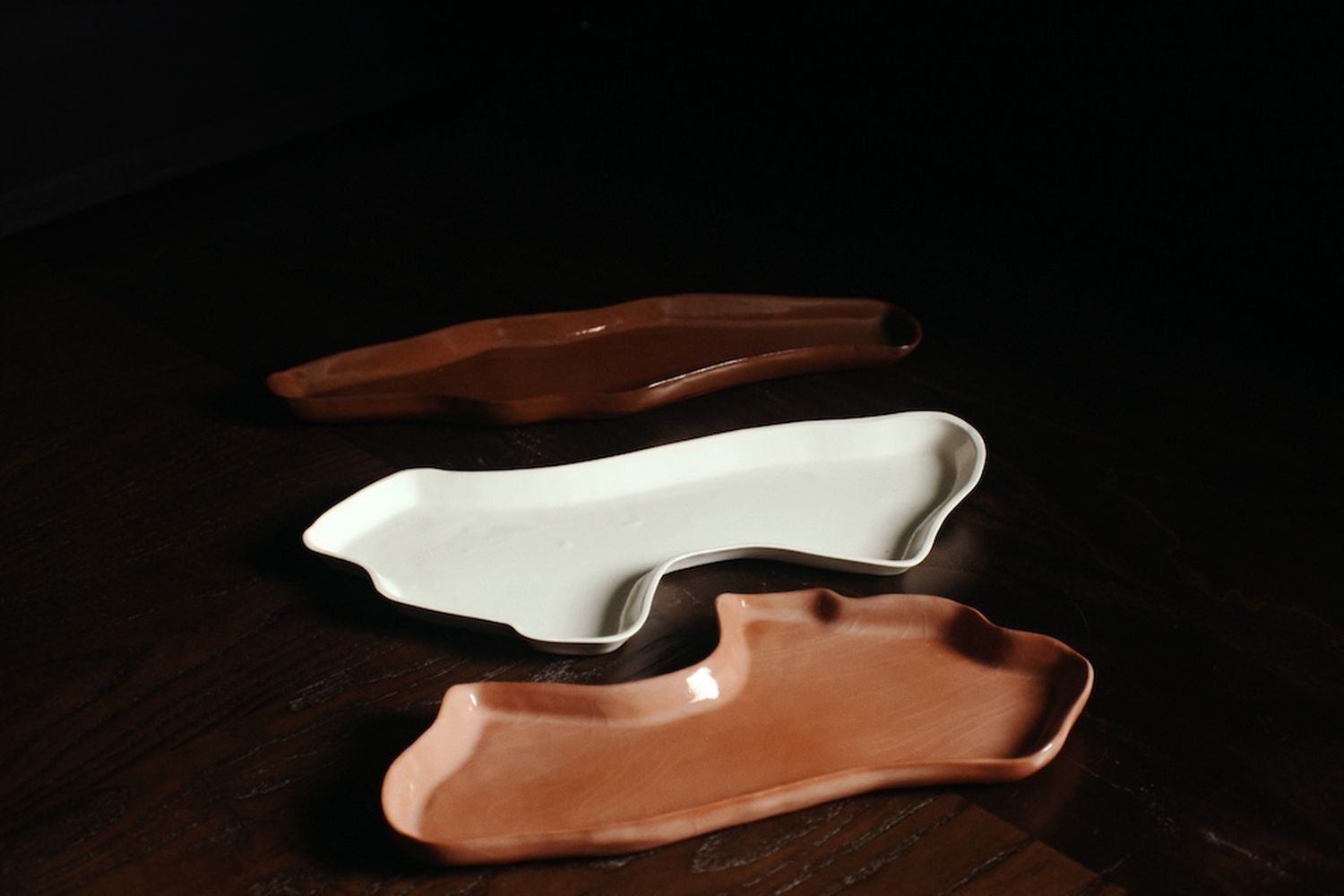 Three abstract ceramic trays in brown, white and tan on a dark wood table.