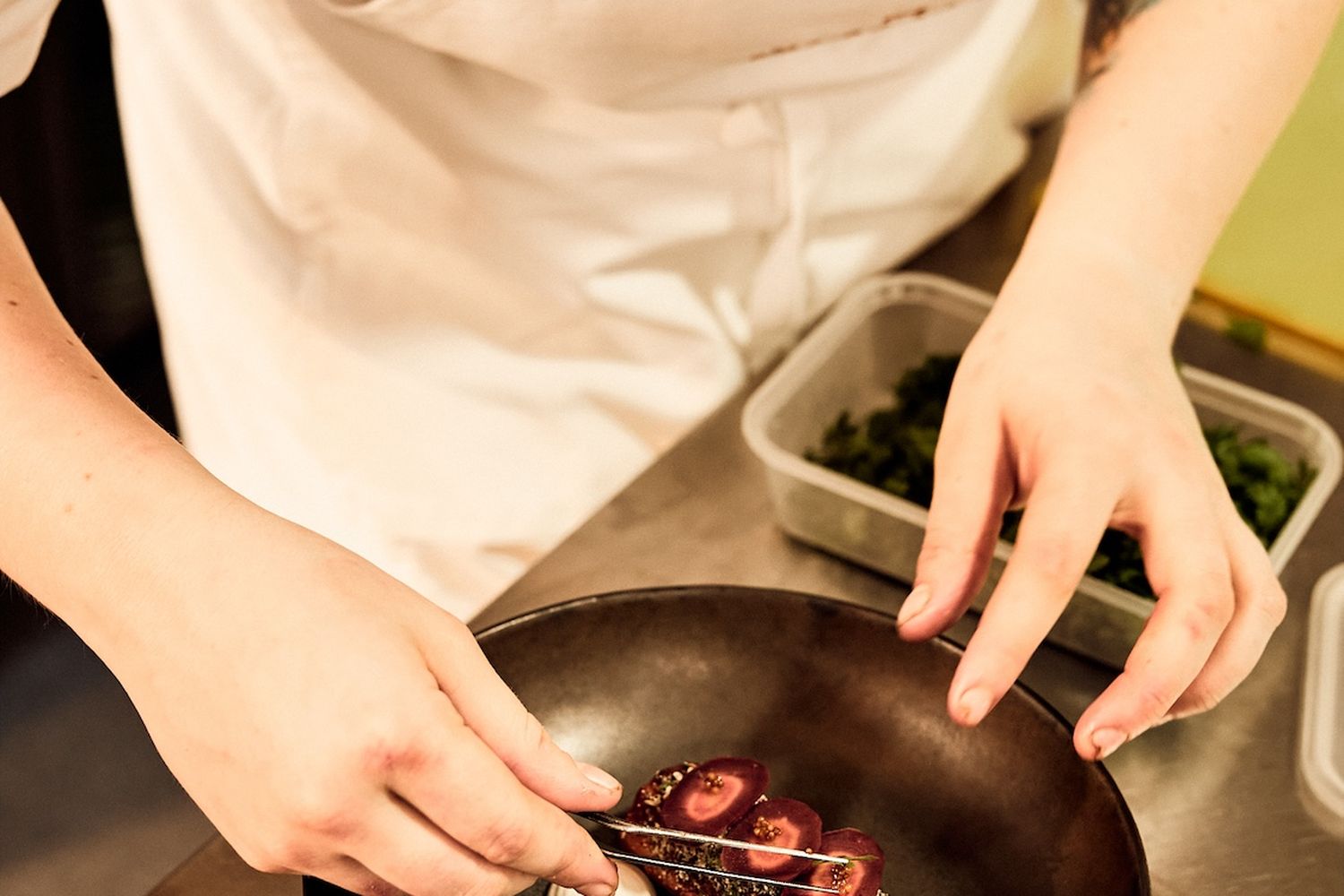 A chef in a white apron is preparing a dish in a black skillet on a stainless steel counter. The dish contains a piece of meat with sauce, garnished with two onions and a cherry tomato. The chef is using a whisk to mix the ingredients together. [G]