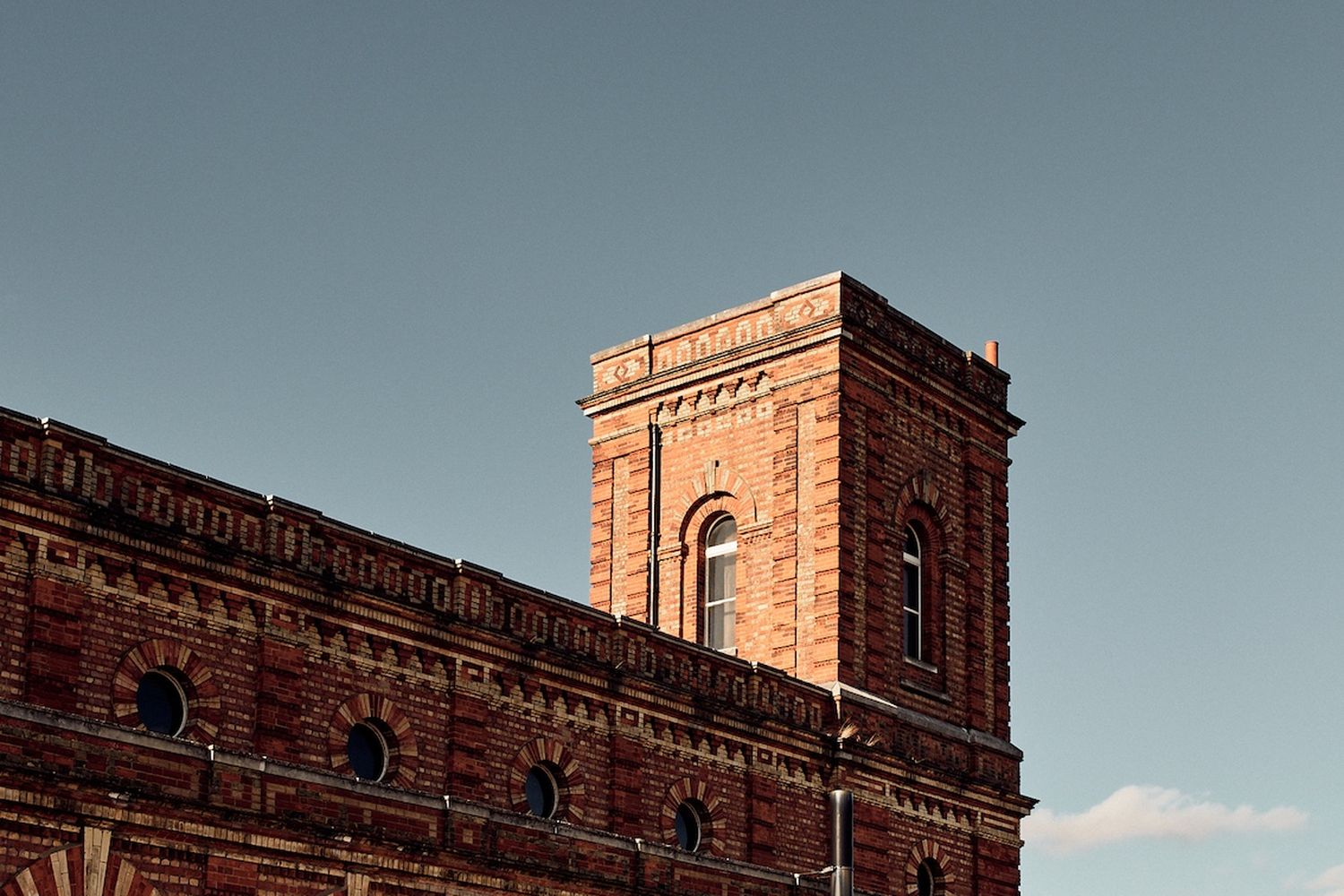 A historic brick building with a tall tower and arched windows stands against a clear blue sky. [G]