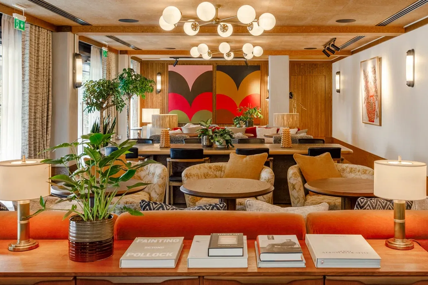The image showcases a cozy and inviting interior space. The room is adorned with a vibrant mural, adding a splash of color to the space. The wooden walls and ceiling create a warm and rustic ambiance. The furniture is well-decorated with various cushions and pillows, providing comfort and style. A wooden table is placed in the center of the room, surrounded by a variety of plants, adding a touch of nature to the space. The room is illuminated by hanging lights, casting a warm glow throughout. The overall atmosphere is one of relaxation and tranquility. [G]