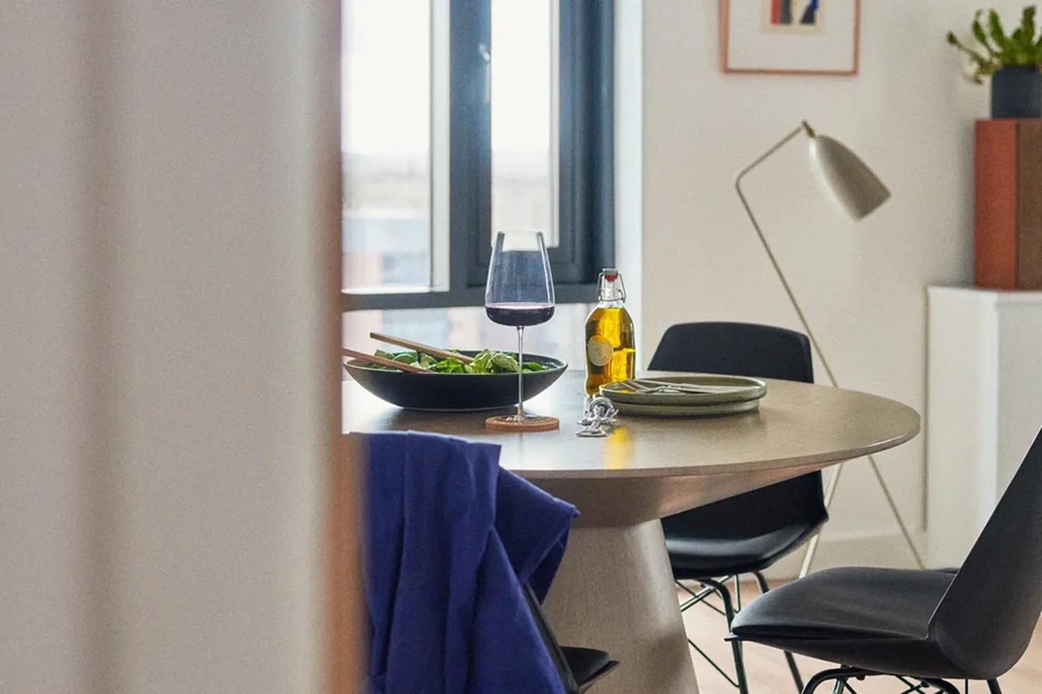 The image features a dining setup with a round table adorned with a bowl of salad, a bottle of beer, and a glass of water. The table is surrounded by chairs, and there's a lamp nearby. The background showcases a window with a view of the cityscape. [G]