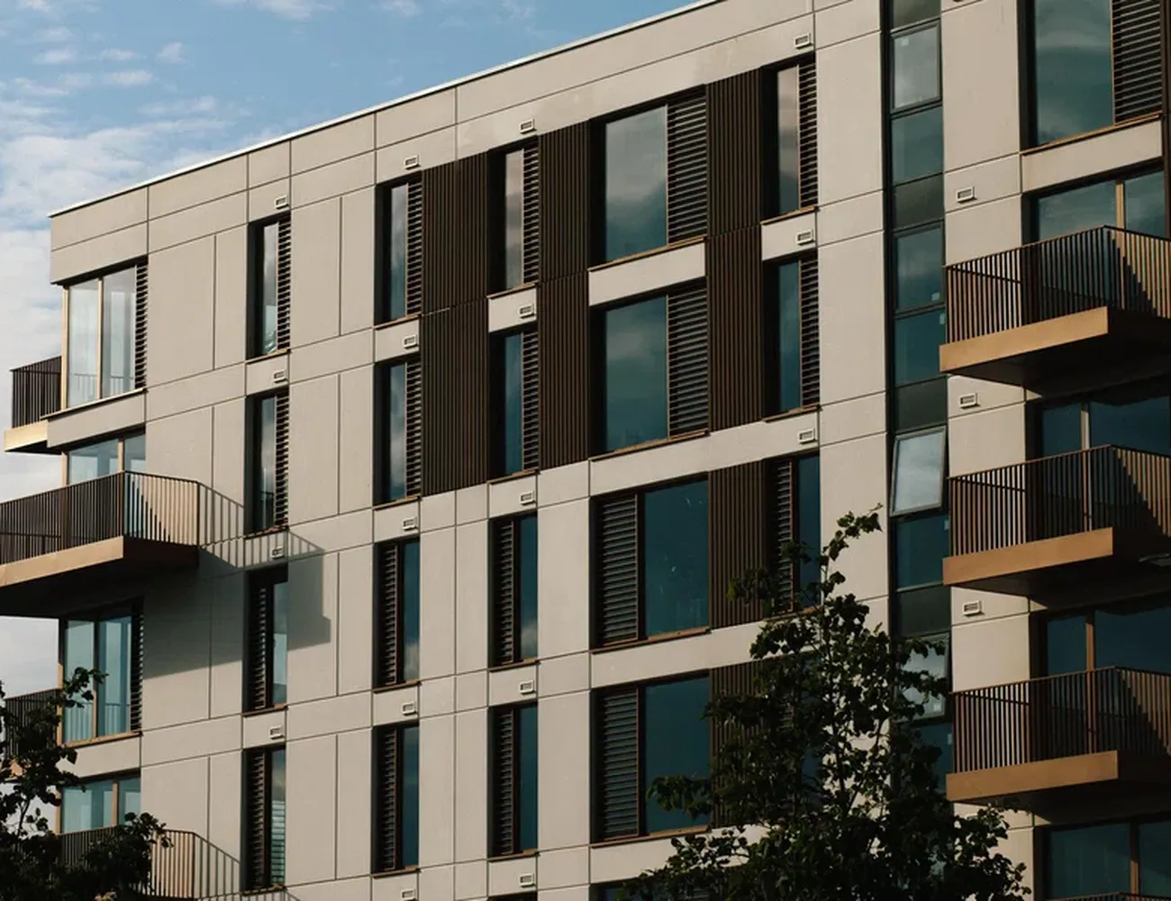 The image showcases a modern apartment building with a unique architectural design. The building features a combination of white and brown panels, creating a harmonious blend. The windows are evenly spaced, with some having brown shutters, adding a touch of warmth to the otherwise cool exterior. The building stands tall against a clear blue sky, with a tree visible in the foreground. The image captures the essence of modern architecture, blending functionality with aesthetics. [G]
