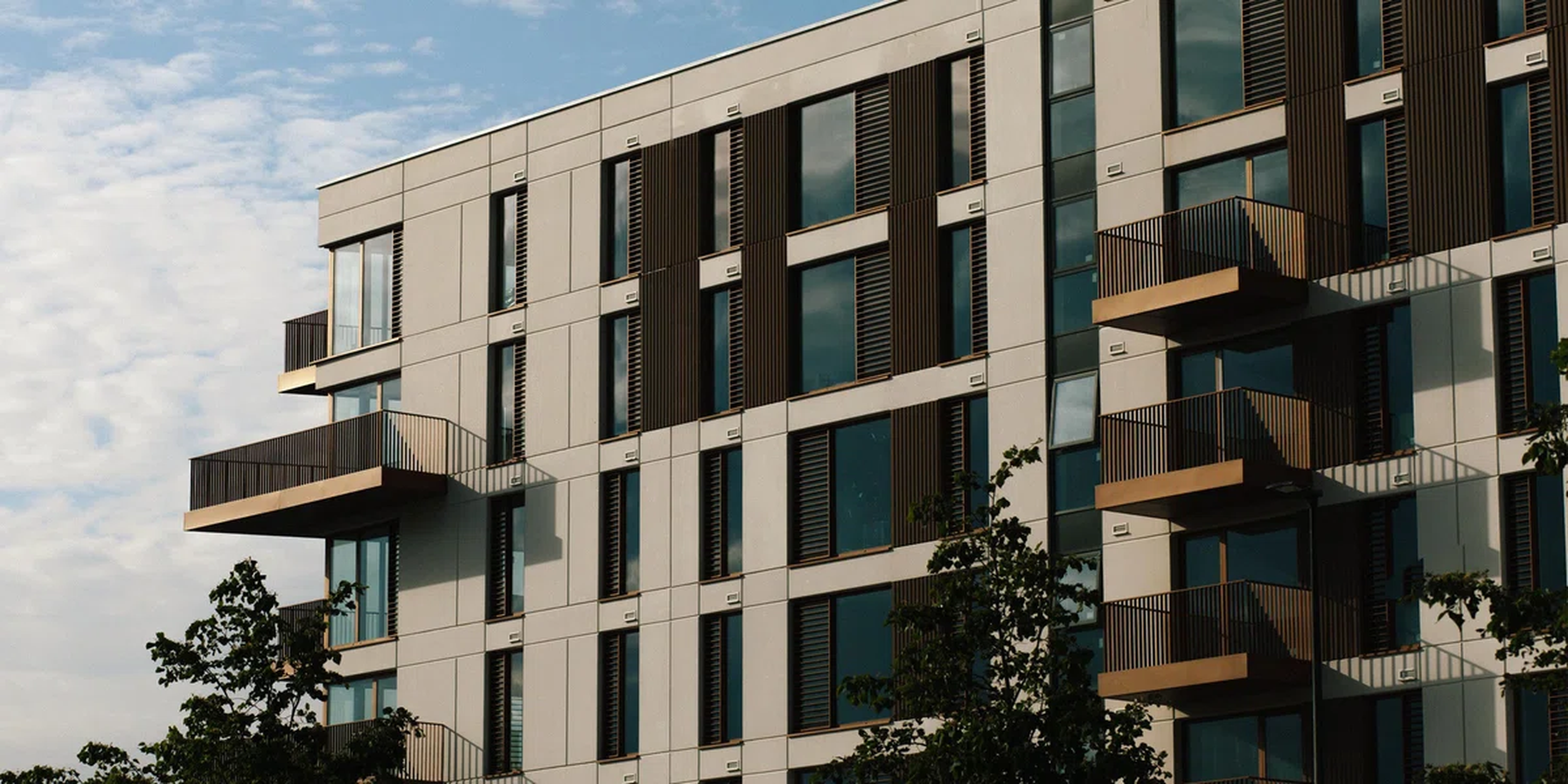 The image showcases a modern apartment building with a unique architectural design. The building features a combination of white and brown panels, creating a harmonious blend. The windows are evenly spaced, with some having brown shutters, adding a touch of warmth to the otherwise cool exterior. The building stands tall against a clear blue sky, with a tree visible in the foreground. The image captures the essence of modern architecture, blending functionality with aesthetics. [G]
