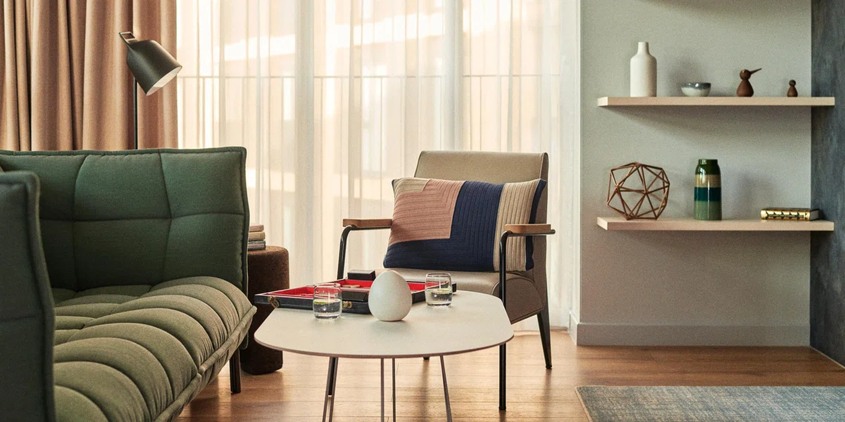 The image features a cozy living room with a beige armchair adorned with a colorful blanket. The armchair is positioned near a white coffee table with a glass of water and a red tray. The room is illuminated by natural light streaming in through white curtains. A green couch is also present in the room. The floor is made of wood, and there's a rug on the floor. The room has a modern aesthetic with a geometric wall and a wooden floor. [G]