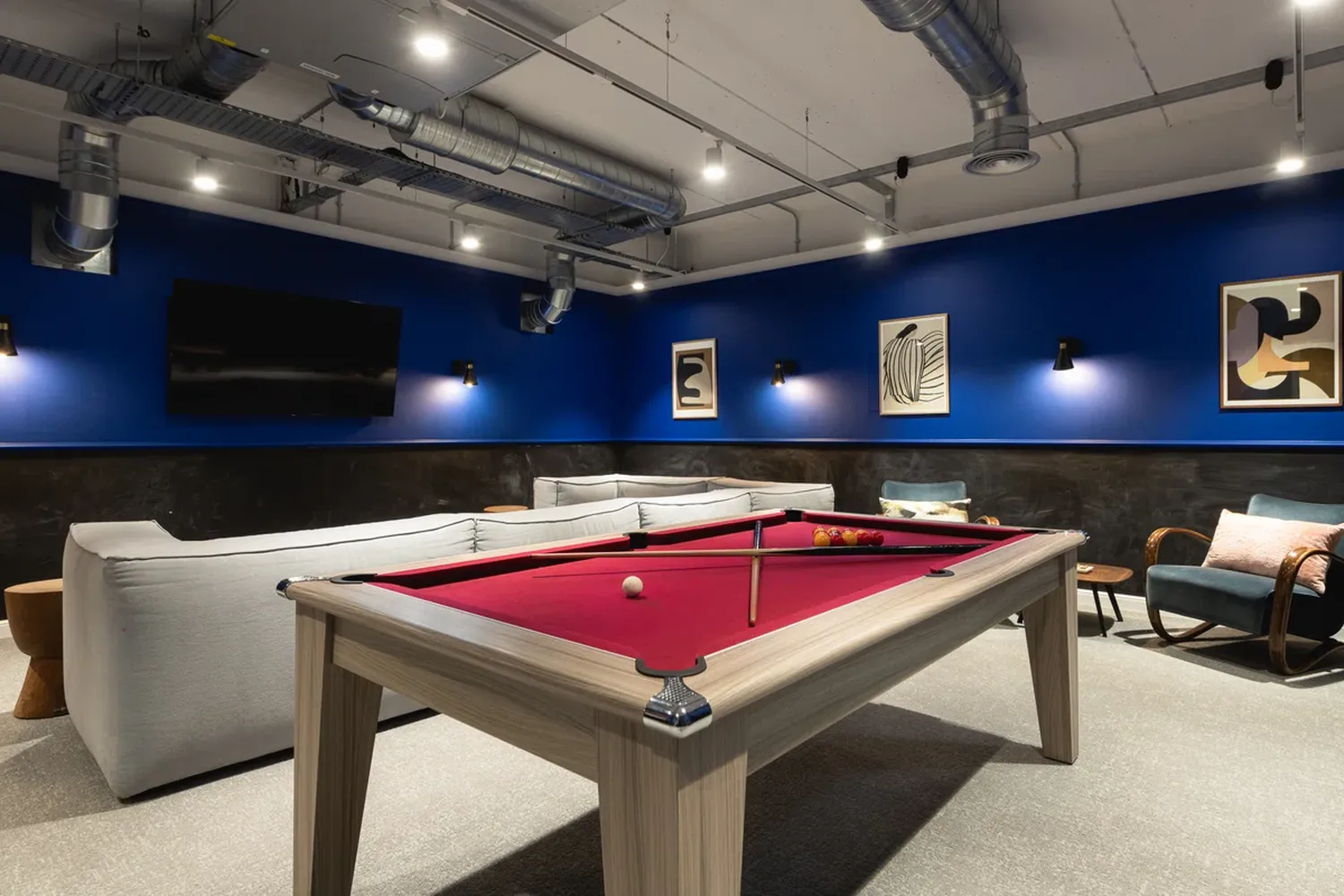 The image features a modern pool table with a vibrant red felt surface, surrounded by a sleek white sofa. The room is illuminated by recessed lighting, casting a warm glow on the objects. Two framed artworks hang on the wall, adding a touch of artistry to the space. The floor is covered in a light gray carpet, providing a neutral backdrop for the objects. The room is well-organized, with each object placed in its designated spot. The overall atmosphere is one of relaxation and leisure, perfect for a game of pool. [G]
