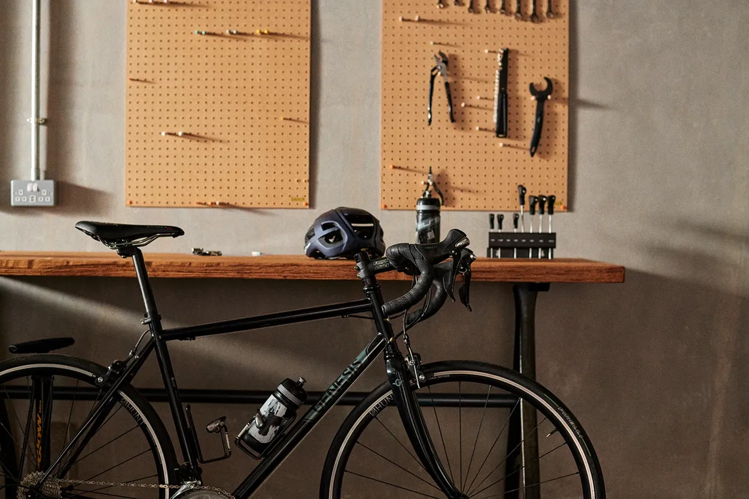 The image features a bicycle parked in a garage or workshop. The bicycle has a helmet on it, indicating it's likely used for commuting or other outdoor activities. The garage or workshop has a pegboard on the wall, which holds various tools and equipment. There are several bolts and wrenches hanging on the pegboard, suggesting the area is used for various tasks such as fixing or working on vehicles. The bicycle is positioned in front of the pegboard, suggesting it's easily accessible for the owner to access the tools. [G]