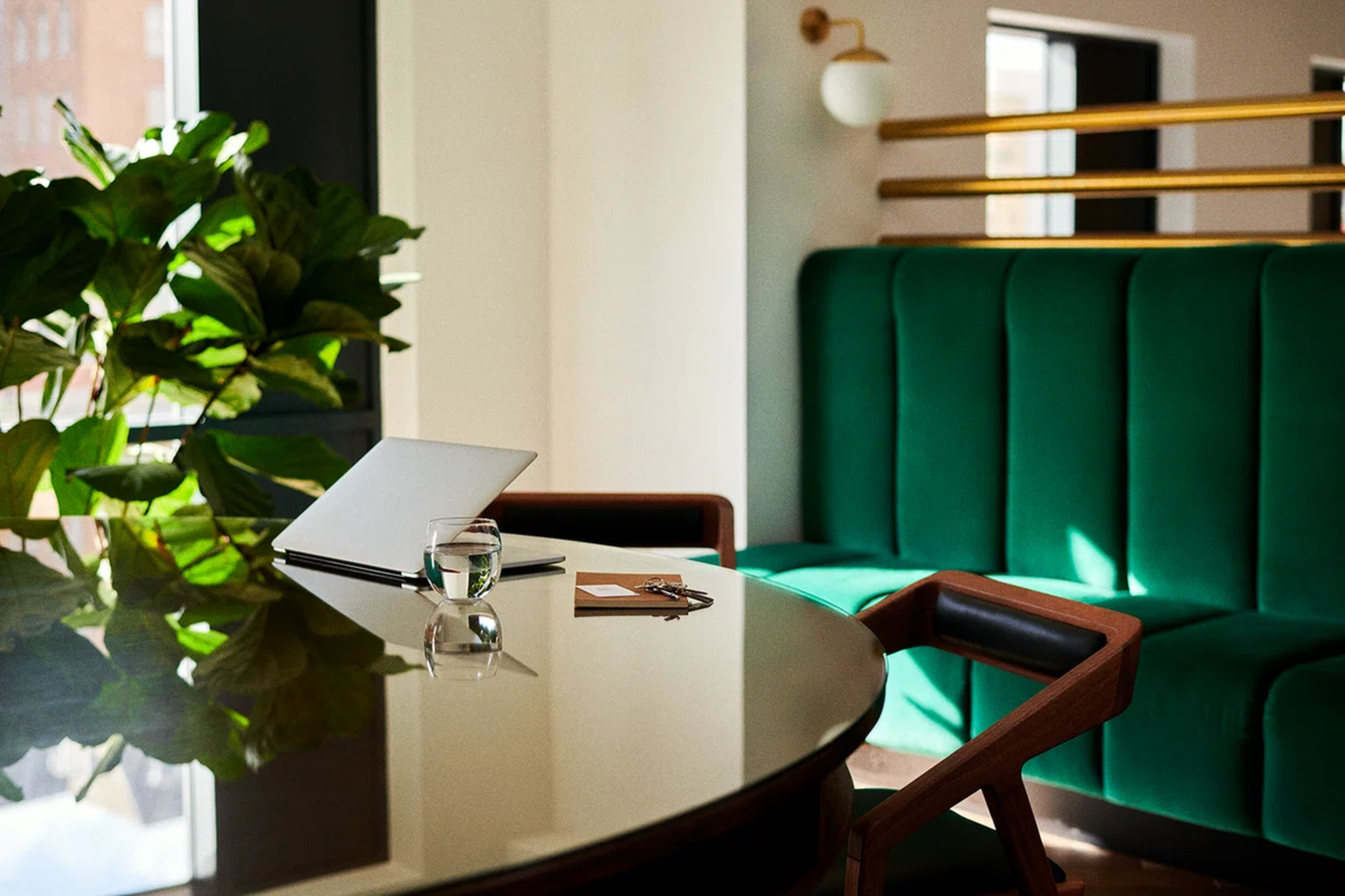 The image features a modern workspace with a sleek black table and a sleek silver laptop. On the table, there are two glasses and a pen. The table is positioned in front of a green sofa with a gold cushion. The background showcases a wall with a white lamp and a plant. The overall atmosphere is calm and focused. [G]