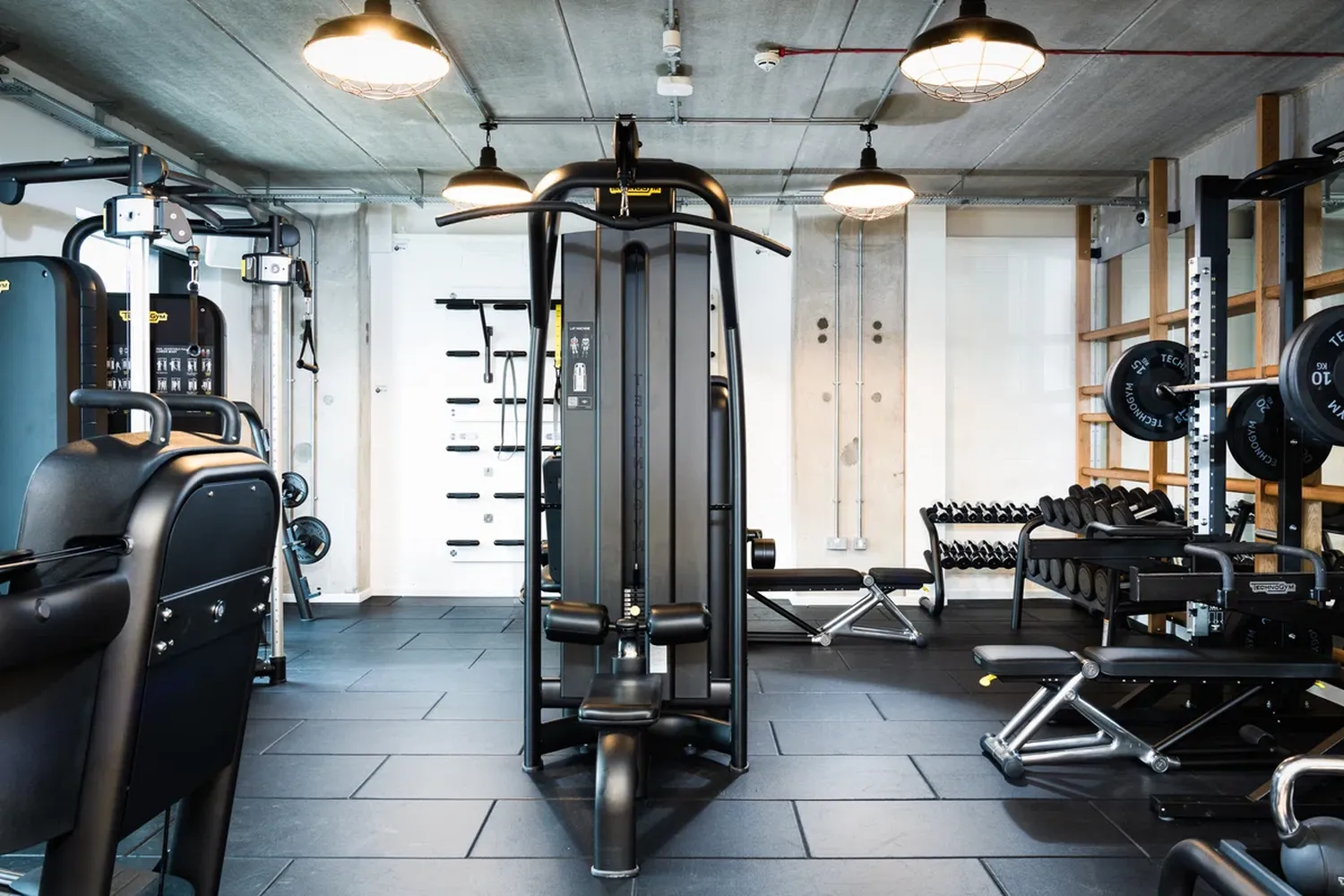The image features a gym with a large black weight machine in the center. The gym has a white wall and a black floor. There are two black exercise machines on the left side of the image. The gym has two hanging lights and a black bench on the right side. The gym is well-lit and spacious. [G]