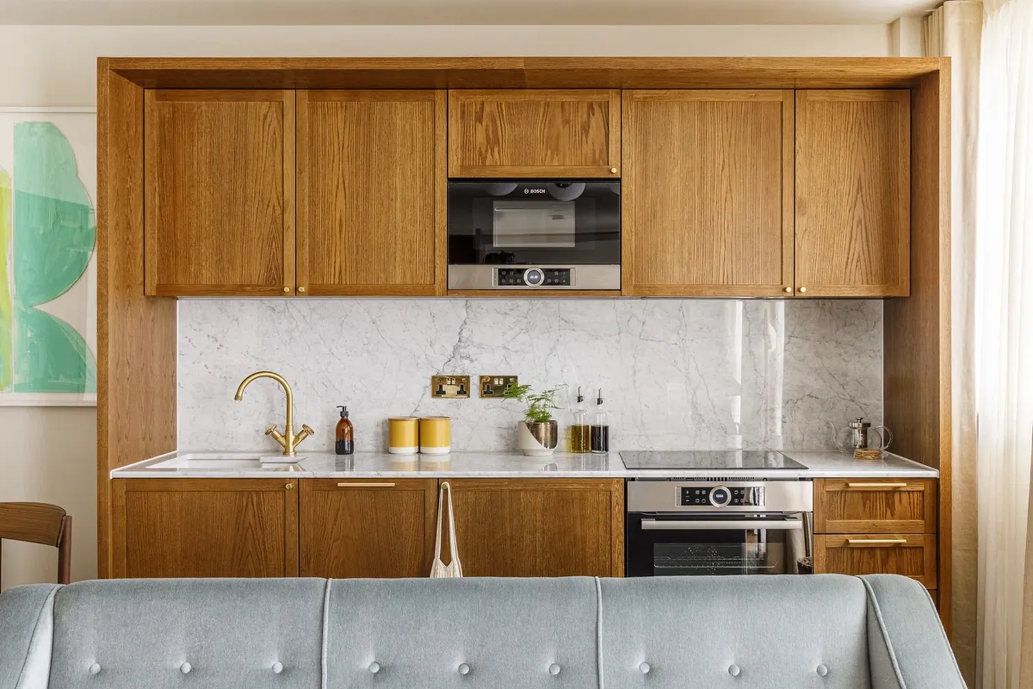 The image showcases a modern kitchen with wooden cabinets and a white marble countertop. The countertop is adorned with various objects such as a gold faucet, a vase with flowers, and a bottle of wine. The cabinets are made of wood and have a sleek design. The kitchen also features a stove with a black oven and a microwave oven. The walls are painted white and have a wooden finish. The overall atmosphere of the kitchen is cozy and inviting. [G]