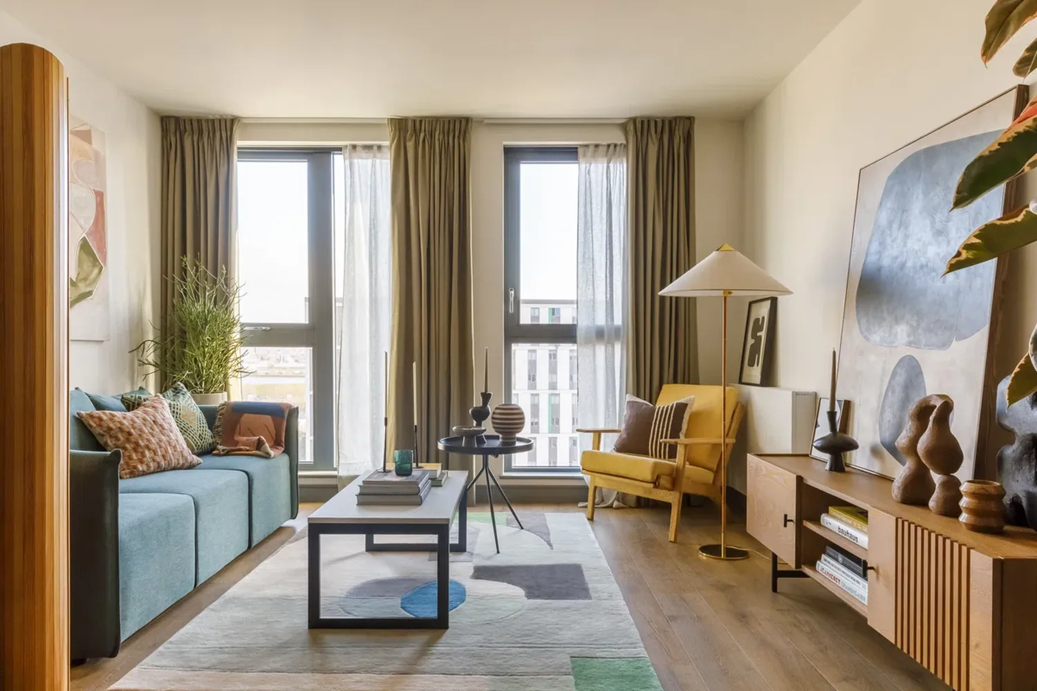 The image features a cozy living room with a soothing color palette. The room is furnished with a blue sofa adorned with colorful pillows, a wooden armchair, and a wooden coffee table. The coffee table is surrounded by a blue rug and a white vase with a green plant. A lamp with a white shade stands on the coffee table, casting a warm glow. The room is illuminated by two large windows, each adorned with beige curtains. The floor is made of wood, and there's a wooden cabinet in the background. The room exudes a sense of comfort and tranquility. [G]