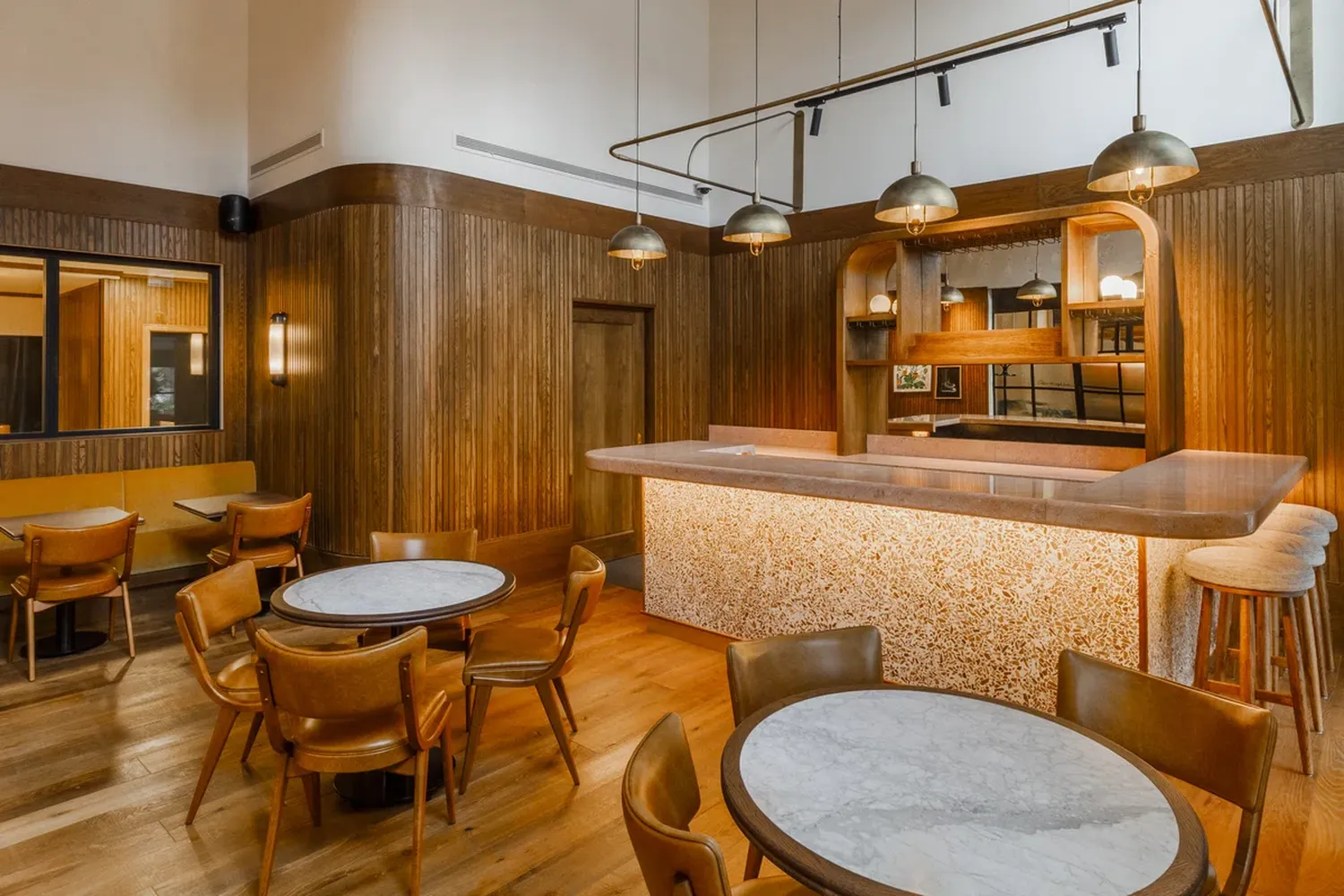 The image features a cozy and inviting interior space. The room is characterized by a warm wooden floor and walls, giving it a rustic charm. The ceiling is adorned with hanging lights, casting a soft glow throughout the space. A large marble counter stands prominently in the center, providing a place for guests to sit and enjoy their meal. The counter is complemented by a wooden door, adding to the room's cozy ambiance. The furniture consists of round tables and chairs, arranged around the counter, inviting guests to sit and enjoy their meal. The overall atmosphere is one of comfort and relaxation, making it an ideal place for social gatherings or relaxation. [G]