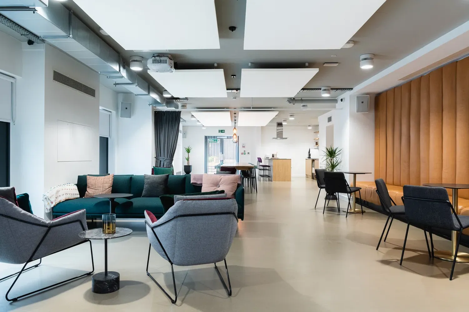 The image features a modern office space with a spacious layout. The room is furnished with a green sofa and a black armchair, both adorned with pillows. The seating arrangement is inviting, with a coffee table in the center. The walls are painted white, creating a bright and airy atmosphere. A large window is visible on the left side of the room, allowing natural light to illuminate the space. A man is present in the background, possibly working or attending a meeting. The room is well-lit, with a ceiling light and a vent for air circulation. The overall design is modern and sleek, making it a suitable workspace. [G]