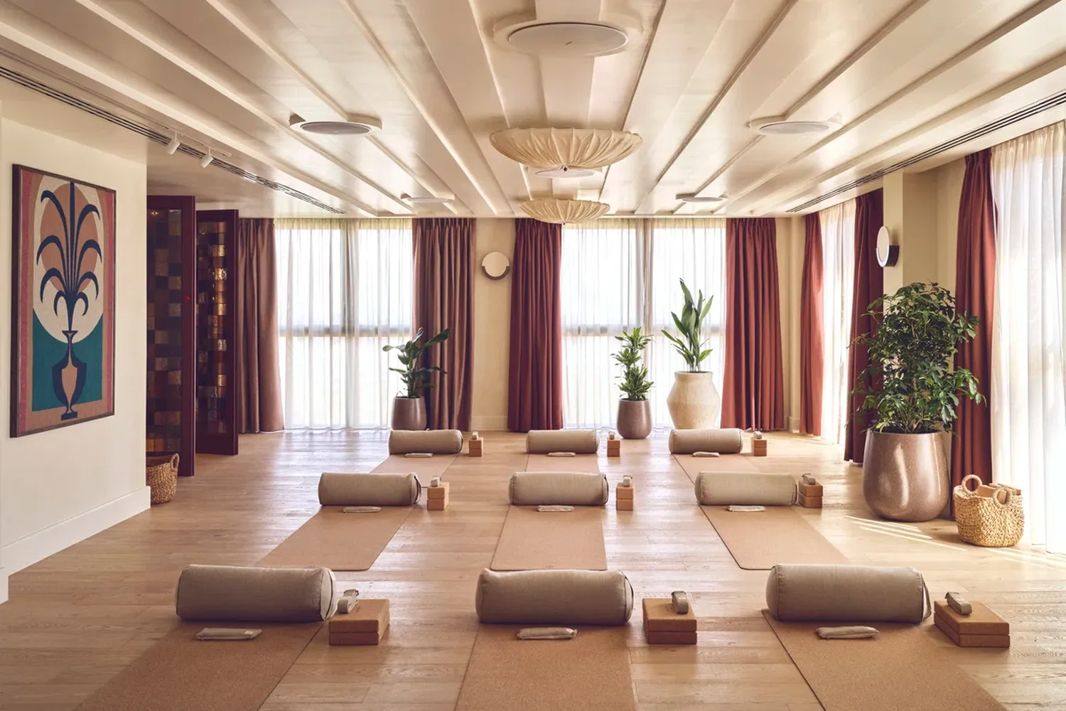 The image depicts a serene yoga room with wooden floors and white walls. The room is furnished with 12 yoga mats arranged in a U-shape, each facing a window. The windows are adorned with red curtains, allowing natural light to filter into the room. The room is illuminated by a ceiling light fixture and a chandelier, creating a warm and inviting atmosphere. The room is well-organized, with each mat and chair neatly placed. The room is empty, suggesting a moment of calm before or after a yoga practice. [G]