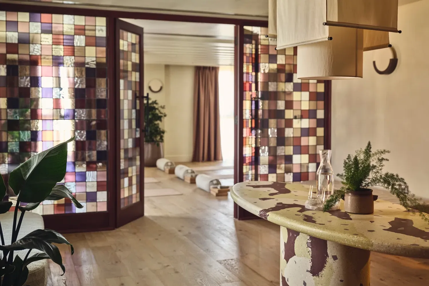 The image showcases a cozy living room with a unique design. The room is adorned with a wooden floor and walls, which are decorated with a checkerboard pattern. A round table with a white top is placed in the center of the room, and on it, there's a vase with a flower. The room also features a large window with a brown curtain, allowing natural light to filter in. A lamp with a white shade is placed on the table, and a plant is placed near the window. The room is well-lit, and the overall ambiance is warm and inviting. [G]
