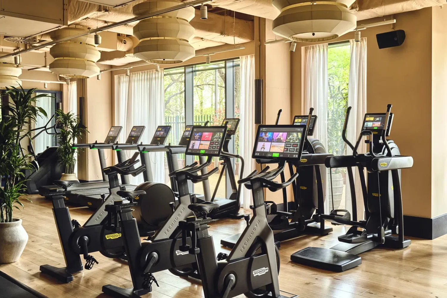 The image showcases a spacious gym room with a variety of exercise equipment. The gym features multiple treadmills, treadmills, and ellipticals, each equipped with a monitor displaying a vibrant app. The gym is well-lit, with hanging light fixtures illuminating the space. The gym is equipped with large windows that offer a view of the outdoors, and the floor is made of polished wood. The gym is designed for both fitness enthusiasts and those looking to maintain a healthy lifestyle. [G]