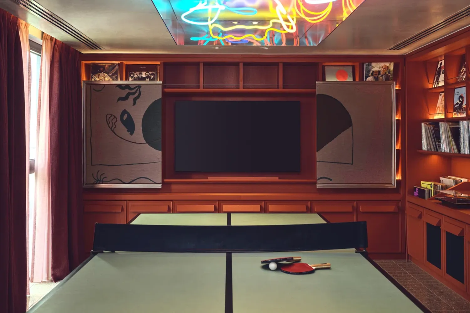 The image features a vibrant and lively entertainment room. Dominating the scene is a large black screen, positioned centrally on the wall. Above it, a neon sign adds a touch of whimsy to the space. To the left of the screen, a table with a ping pong table is set up, ready for a game. The table is adorned with a red paddle and a white ball, hinting at the fun-filled atmosphere of the room. The walls are adorned with various posters and pictures, adding to the room's character. The room is illuminated by a neon light, casting a warm glow that enhances the overall ambiance. [G]