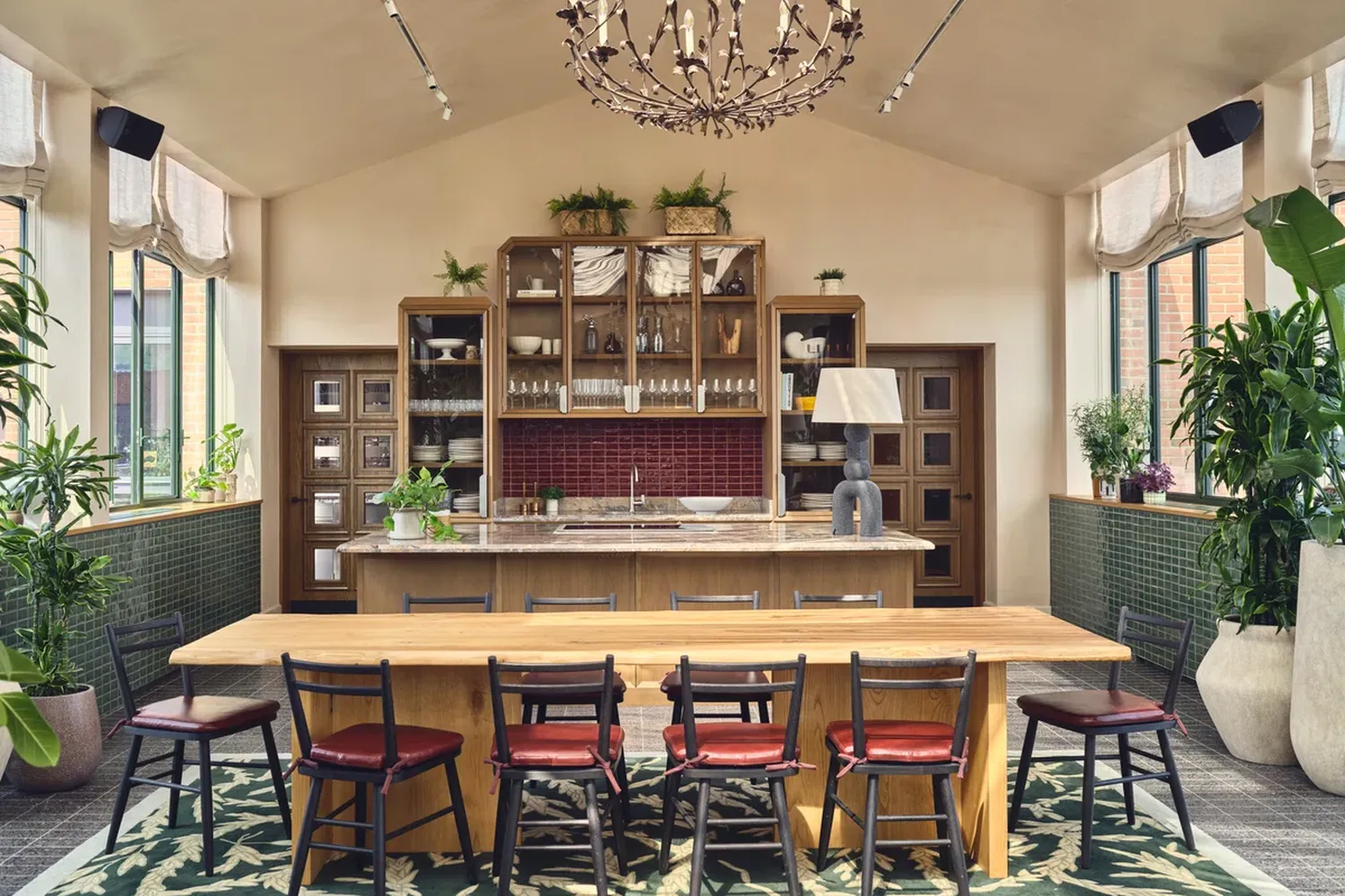 The image showcases a sophisticated kitchen with a wooden counter and a chandelier hanging from the ceiling. The counter is adorned with various objects such as a vase, a lamp, and a vase with flowers. The kitchen features a wooden cabinet with glass doors, and there are several chairs around the counter. The floor is covered with a green rug. The room is well-lit, and there are plants placed in the cabinets. The overall ambiance is elegant and inviting. [G]