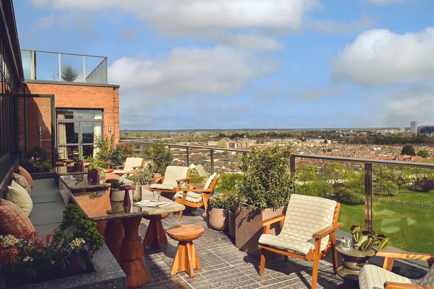 The image captures a serene rooftop patio, offering a panoramic view of the cityscape. The patio is furnished with wooden furniture and chairs, inviting relaxation. The patio is surrounded by potted plants and flowers, adding a touch of nature to the urban setting. The patio is elevated, offering a panoramic view of the city below. The image is taken from a high vantage point, providing a comprehensive view of the cityscape. The overall scene is peaceful and tranquil, making it a perfect spot for a peaceful afternoon. [G]