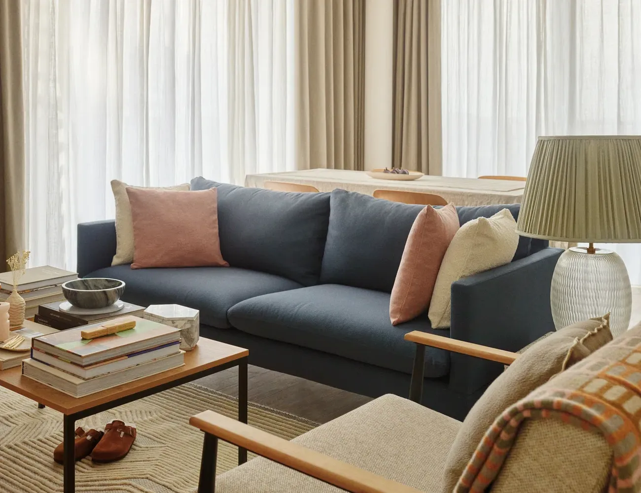 The image showcases a cozy living room with a blue sofa adorned with pink and white pillows. The sofa is positioned in front of a wooden coffee table with a bowl of food and a stack of books. To the left of the sofa, there's a wooden chair with a lamp on it. The room is illuminated by natural light from a window with white curtains. The floor is covered with a rug, and a pair of flip-flops can be seen on the floor. The overall atmosphere is warm and inviting. [G]