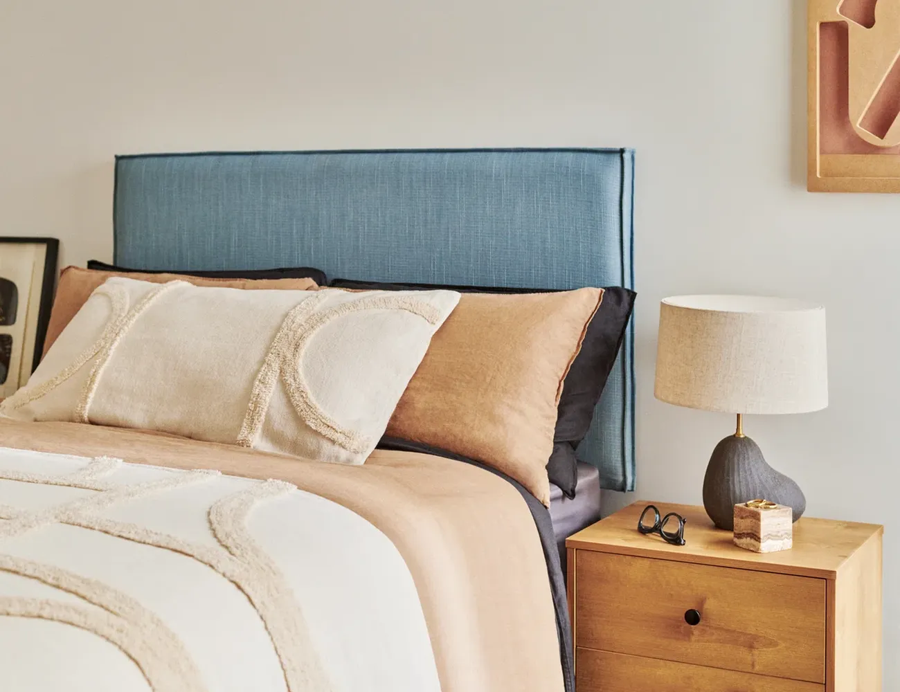 The bedroom features a blue headboard, a bed with beige and brown pillows, and a wooden nightstand with a lamp and sunglasses. [G]