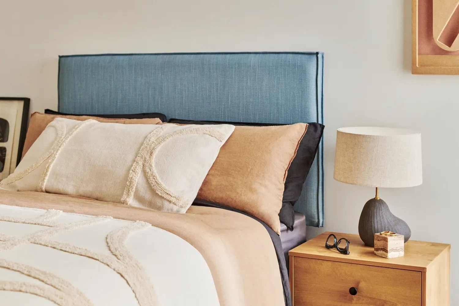 The bedroom features a blue headboard, a bed with beige and brown pillows, and a wooden nightstand with a lamp and sunglasses. [G]