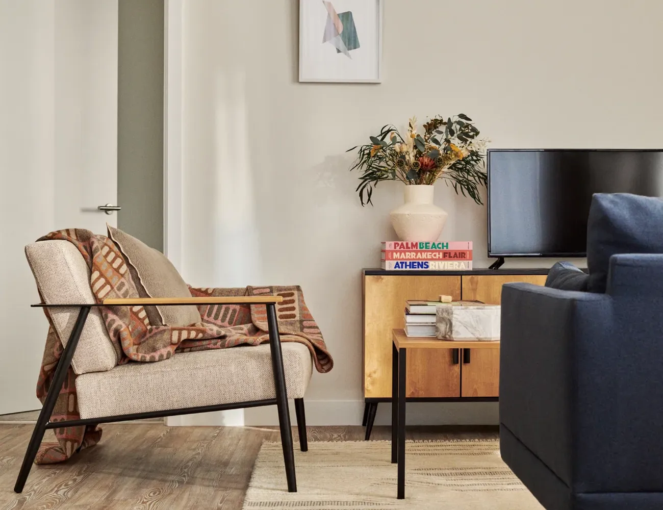 The image showcases a cozy living room with a beige sofa adorned with a brown and white striped blanket. Adjacent to the sofa, a wooden coffee table holds a stack of books and a vase with a green plant. A black television is mounted on the wall, and a white door is visible on the right side of the image. The room is decorated with a white wall, a blue armchair, and a wooden cabinet with a pink vase. The arrangement of objects creates a harmonious and inviting atmosphere. [G]