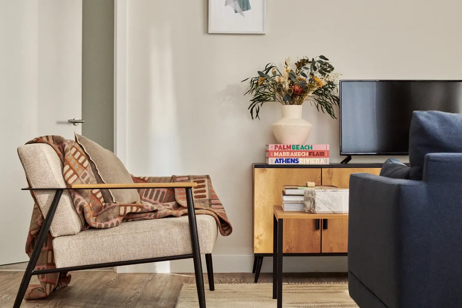 The image showcases a cozy living room with a beige sofa adorned with a brown and white striped blanket. Adjacent to the sofa, a wooden coffee table holds a stack of books and a vase with a green plant. A black television is mounted on the wall, and a white door is visible on the right side of the image. The room is decorated with a white wall, a blue armchair, and a wooden cabinet with a pink vase. The arrangement of objects creates a harmonious and inviting atmosphere. [G]