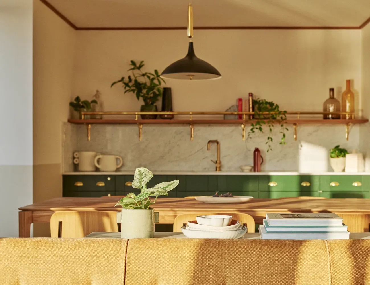 The kitchen is a cozy space with a wooden table and a green countertop. On the table, there are white plates, a green plant, and a white mug. Above the countertop, there's a gold faucet. The kitchen has a wooden cabinet with a gold handle and a green countertop. The walls are painted in a light color, and there are plants in the cabinets. [G]