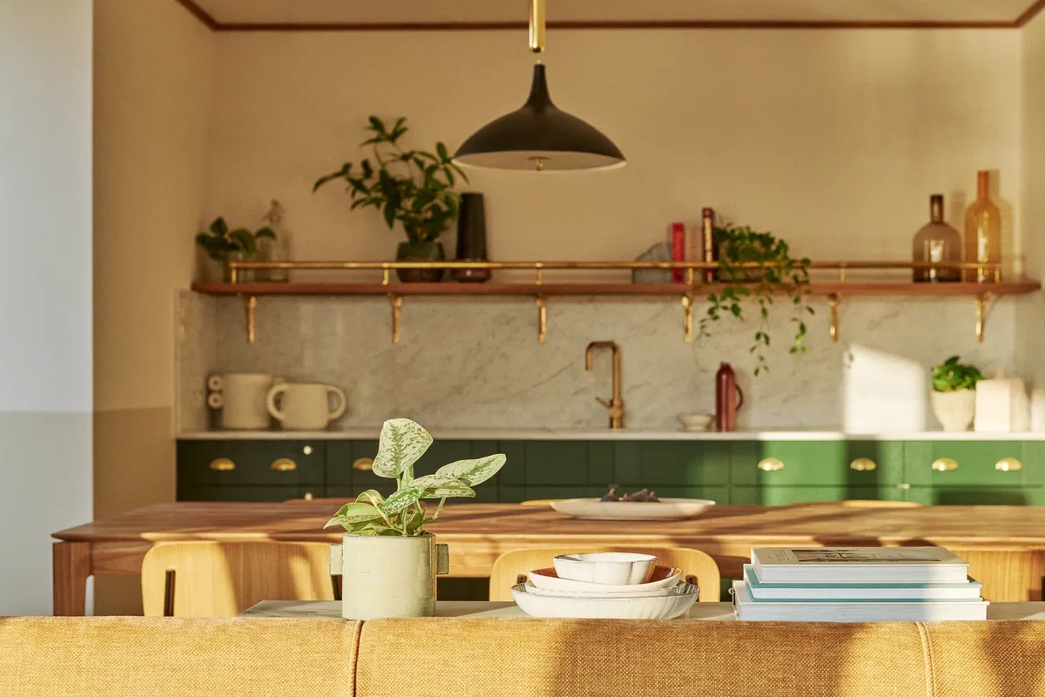 The kitchen is a cozy space with a wooden table and a green countertop. On the table, there are white plates, a green plant, and a white mug. Above the countertop, there's a gold faucet. The kitchen has a wooden cabinet with a gold handle and a green countertop. The walls are painted in a light color, and there are plants in the cabinets. [G]