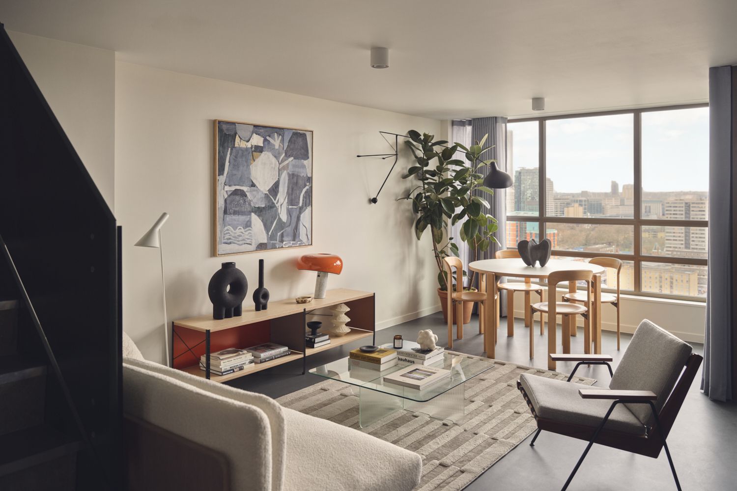 A modern living room with a glass coffee table, a white sofa, and a wooden dining table. The room has a large window overlooking a cityscape. The room features a large abstract painting, a vase, a lamp, and a plant. The room also has a rug and a rug runner. [G]