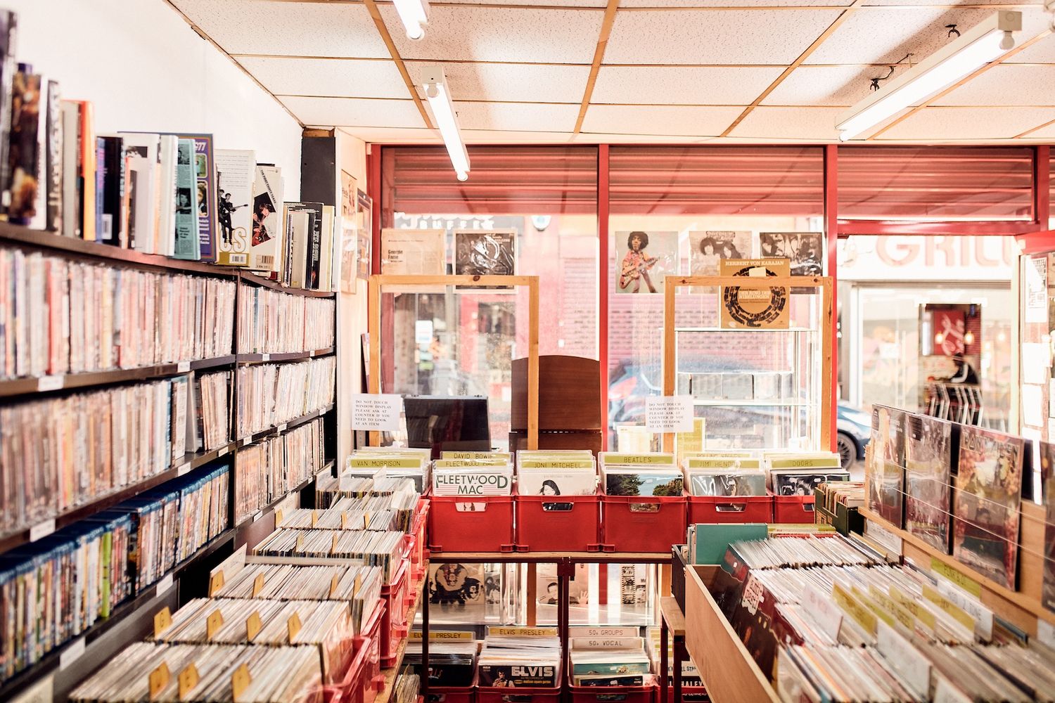 The image features a store filled with a variety of CDs and vinyl records. The store is well-organized with shelves filled with different types of records, including albums, singles, and compilations. The store is located in a building with a red door and a window that offers a view of the outside. The store is well-lit, with a clock hanging on the wall. The store is a music lover's paradise, offering a wide selection of music for all tastes. [G]