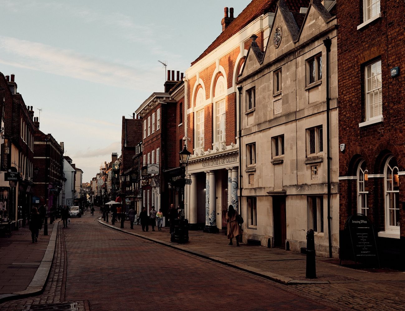 A quaint town street lined with historic buildings, people strolling, and a car parked on the side. [G]