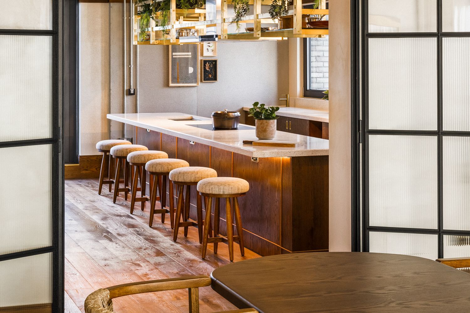 A cozy bar with a wooden counter and stools, surrounded by plants and a window. [G]