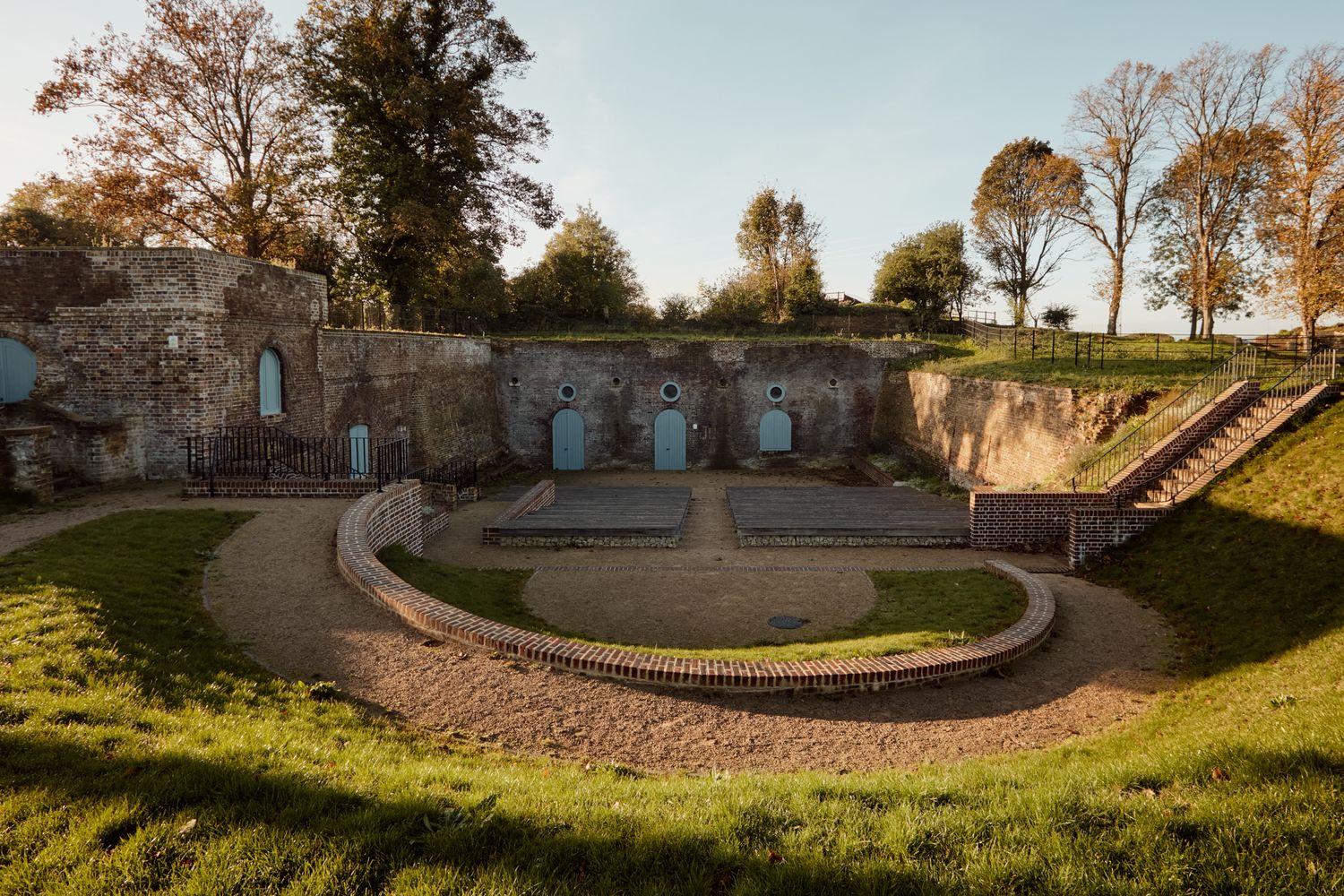 The image captures the historic Fort of Kent, a UNESCO World Heritage Site in Kent, England. The scene is set against a backdrop of lush greenery, with a brick wall encircling the courtyard. The courtyard is a circular space, with four blue doors and four brick columns. The walls of the courtyard are made of stone, and the ground is covered in grass. The overall atmosphere is serene and peaceful, with the sun casting a warm glow on the scene. [G]