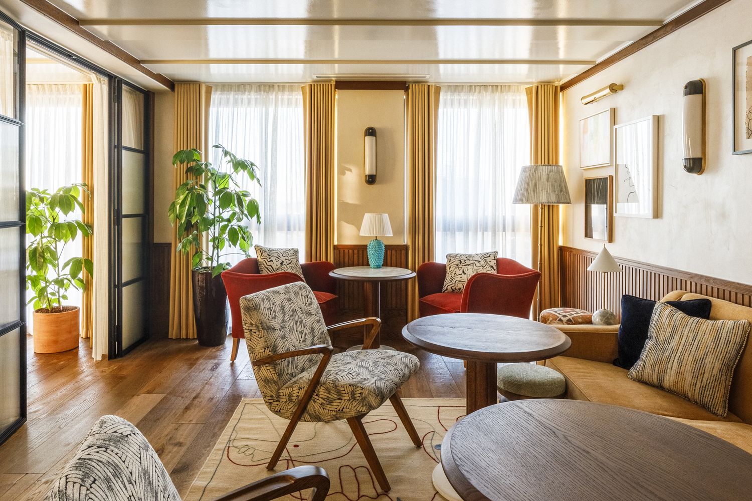 A cozy living room with a wooden floor, a red armchair, a white and gold patterned chair, a round wooden table, a blue vase, a potted plant, and a wooden floor lamp. [G]