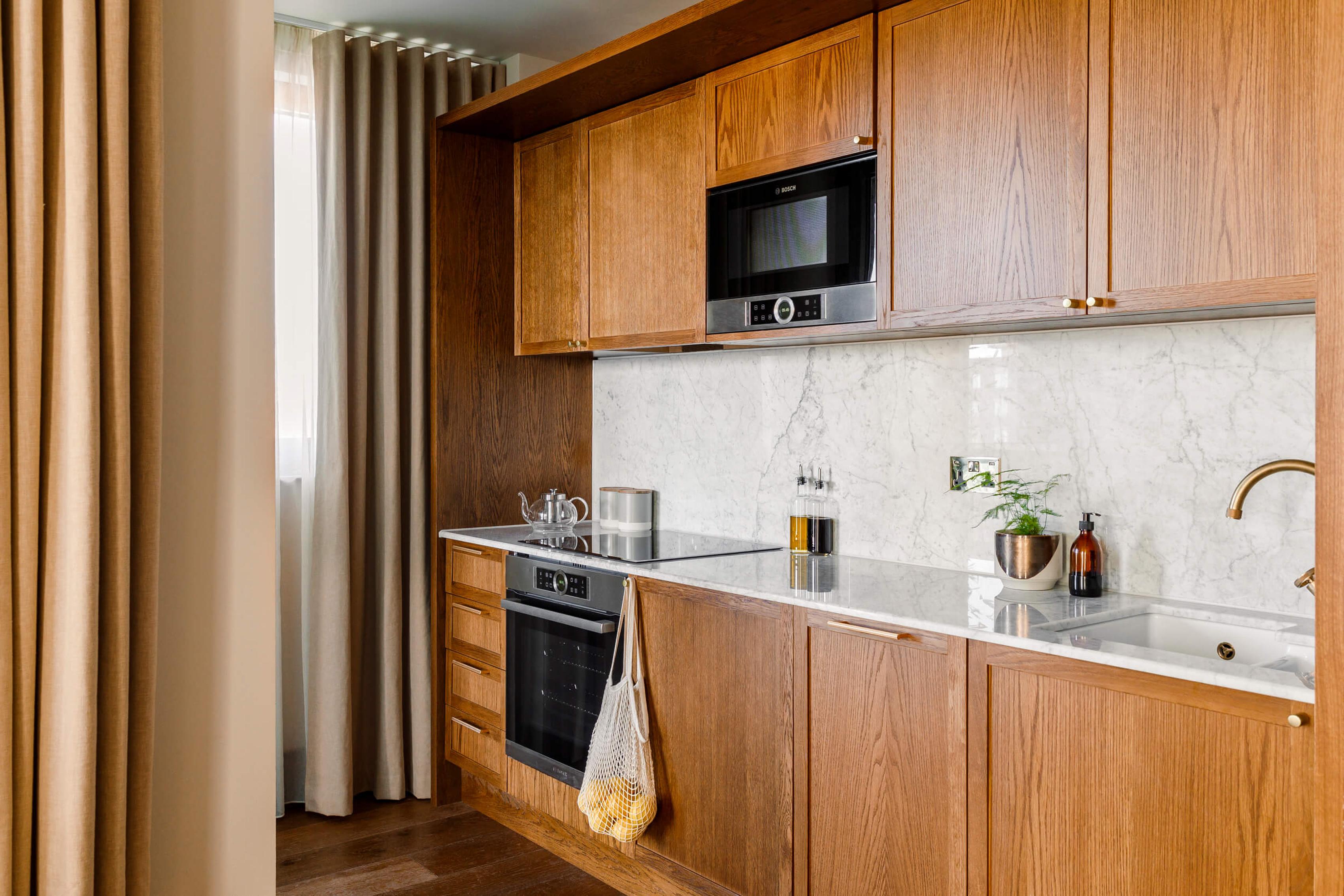 The image features a modern kitchen with wooden cabinets and a white marble countertop. A microwave oven is placed above the countertop, and a sink is situated below it. A window with a curtain is visible on the left side of the image. A hanging plant is also present in the kitchen. [G]