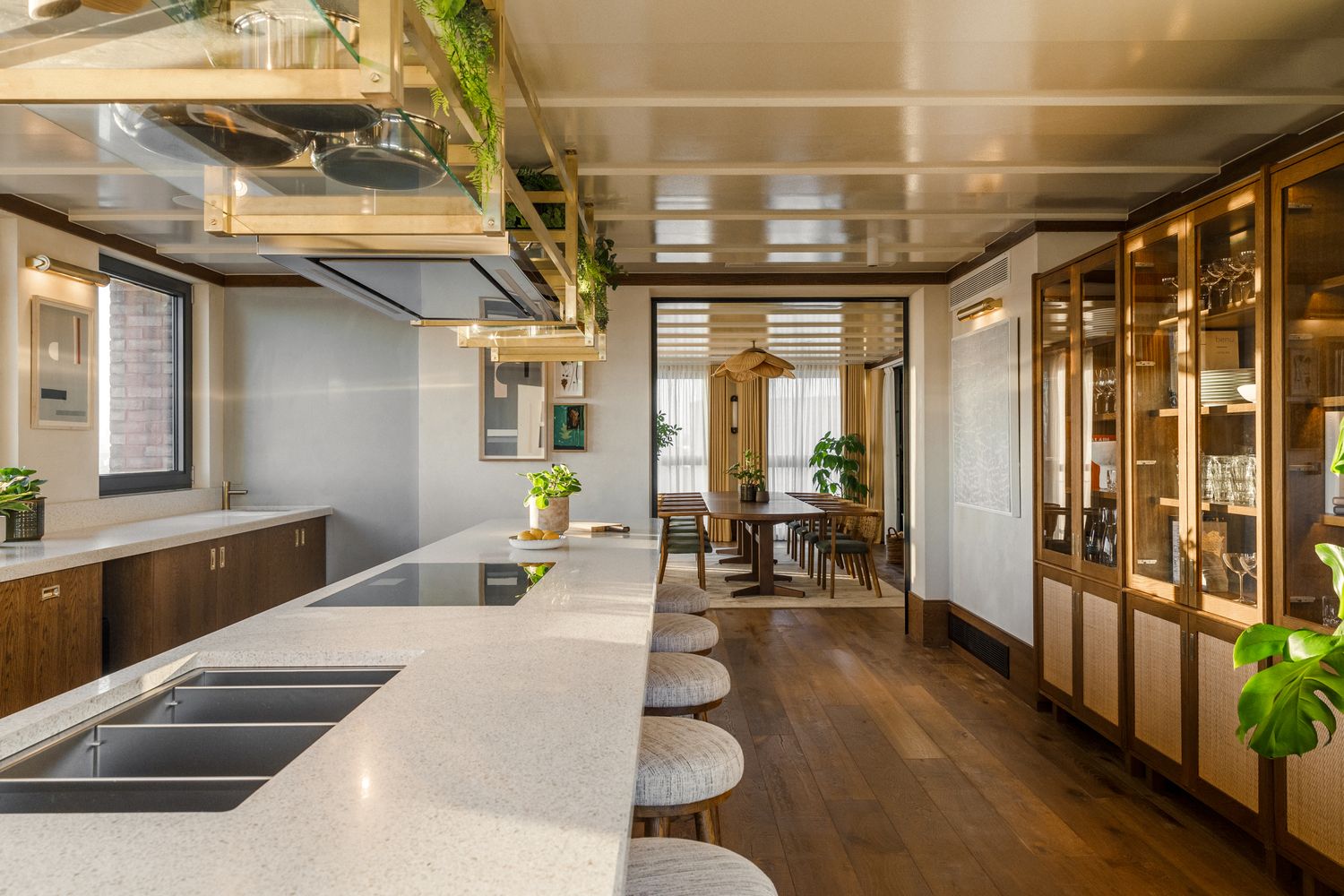 A modern kitchen with a white island counter, stainless steel hood, and pendant lights. The island has a white countertop and a wooden table with a plant. The kitchen has a wooden floor and a wooden ceiling. The room has a window with white curtains and a plant on the window sill. [G]